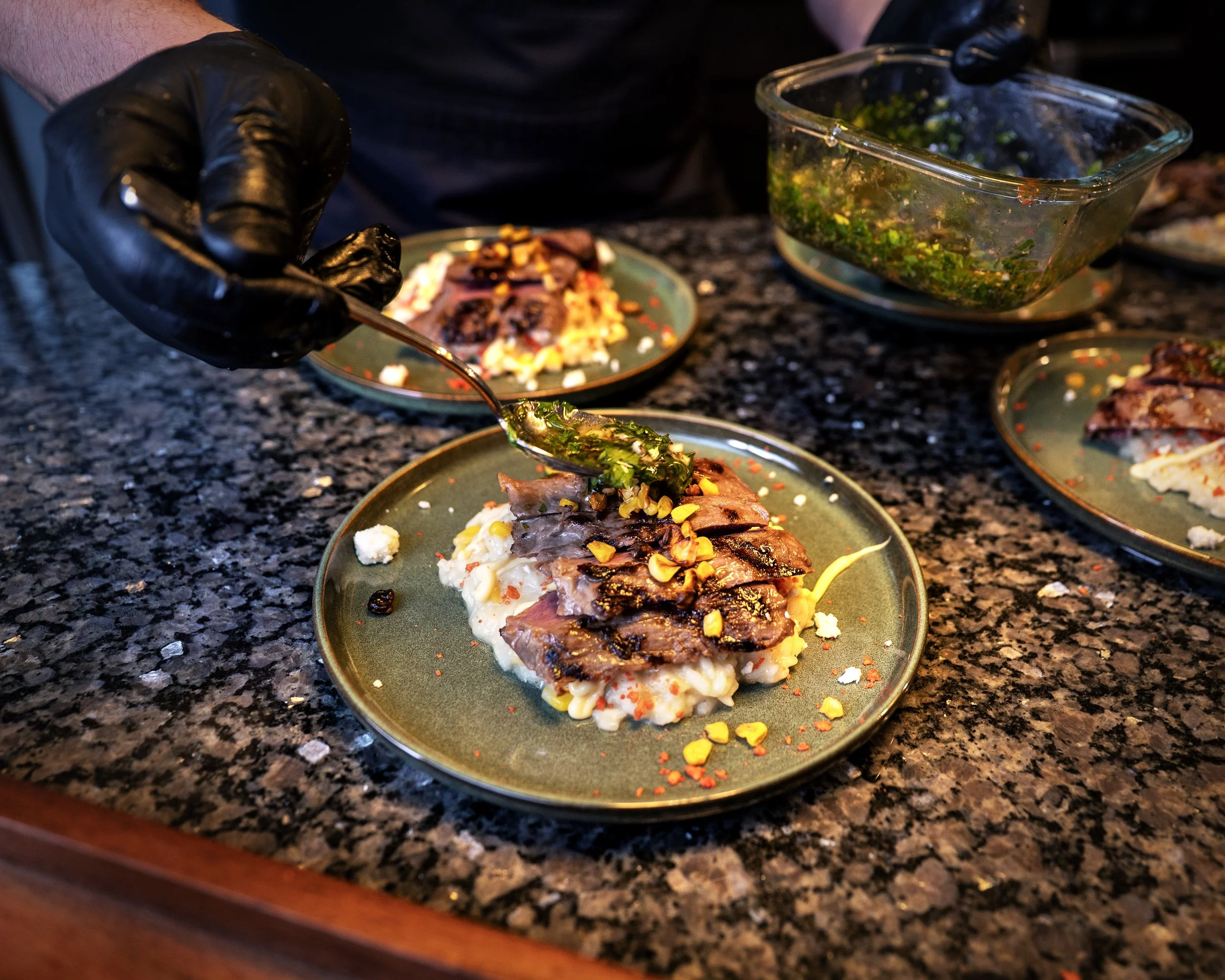 Close-up of a plated meal with grilled fish topped with herbs and spices, garnished with colorful seasoning, on a green plate on a granite countertop. In the background, a person wearing black gloves is preparing more food around a glass dish of chop