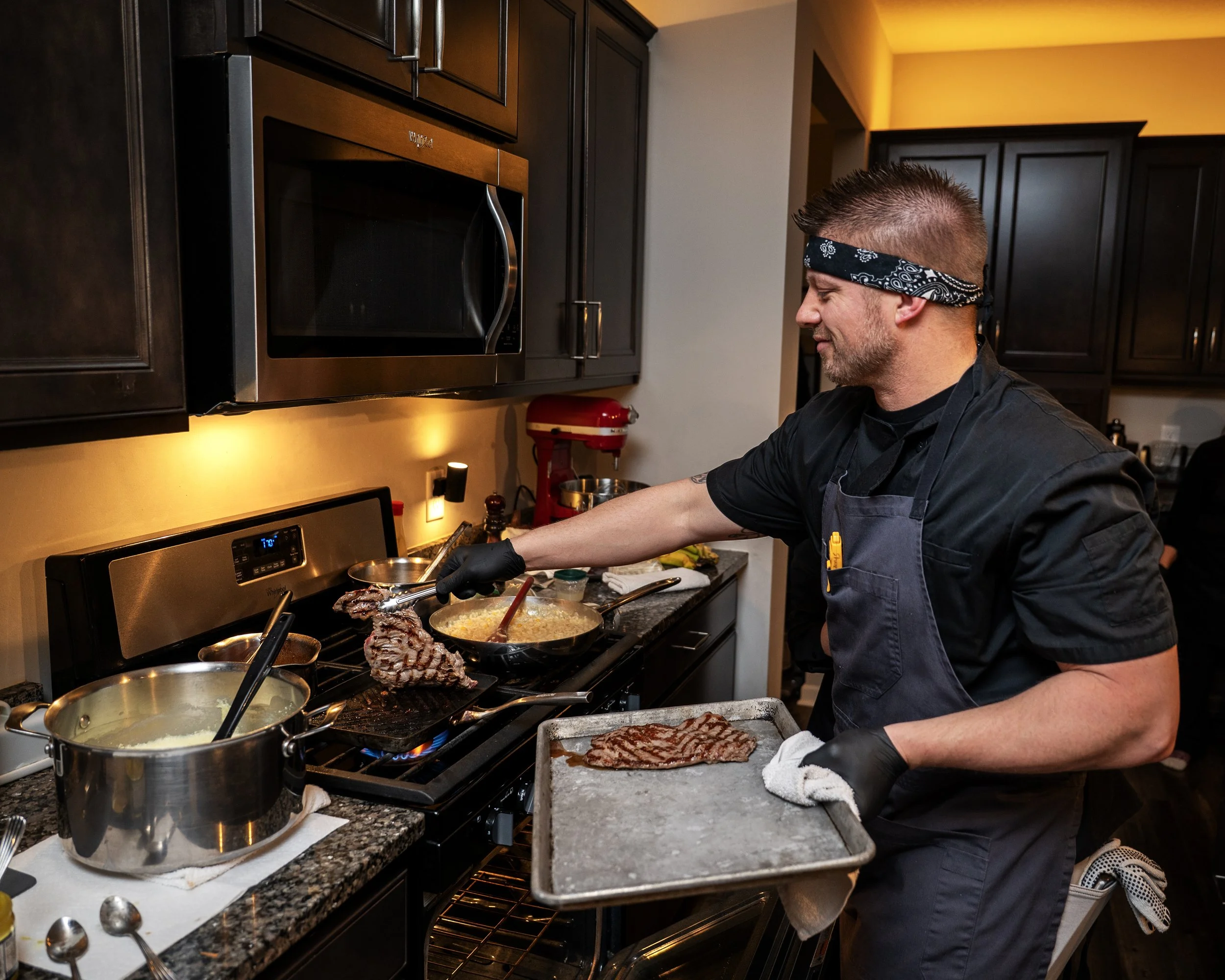 A man with a black bandana and black apron cooking meat on a stovetop in a kitchen. The stove has various pots and pans, and the man is holding a baking sheet. Dark wooden cabinets and a microwave are visible in the background.
