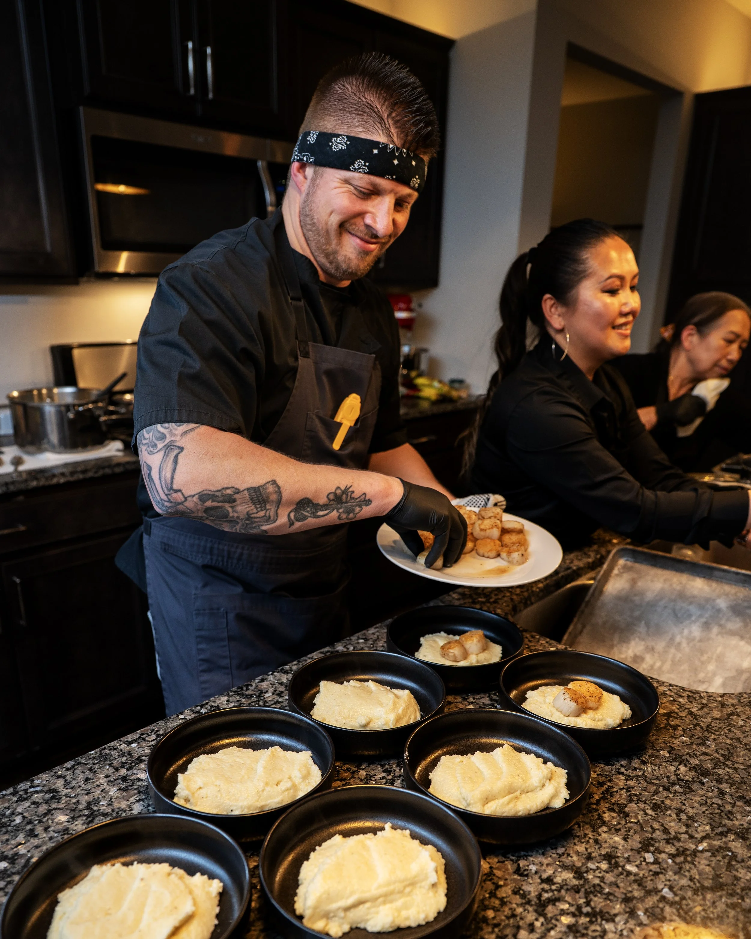 A person with tattoos on the arm, wearing a black bandana and black apron, serving mashed potatoes on black plates in a kitchen. Other people are present in the background, smiling.