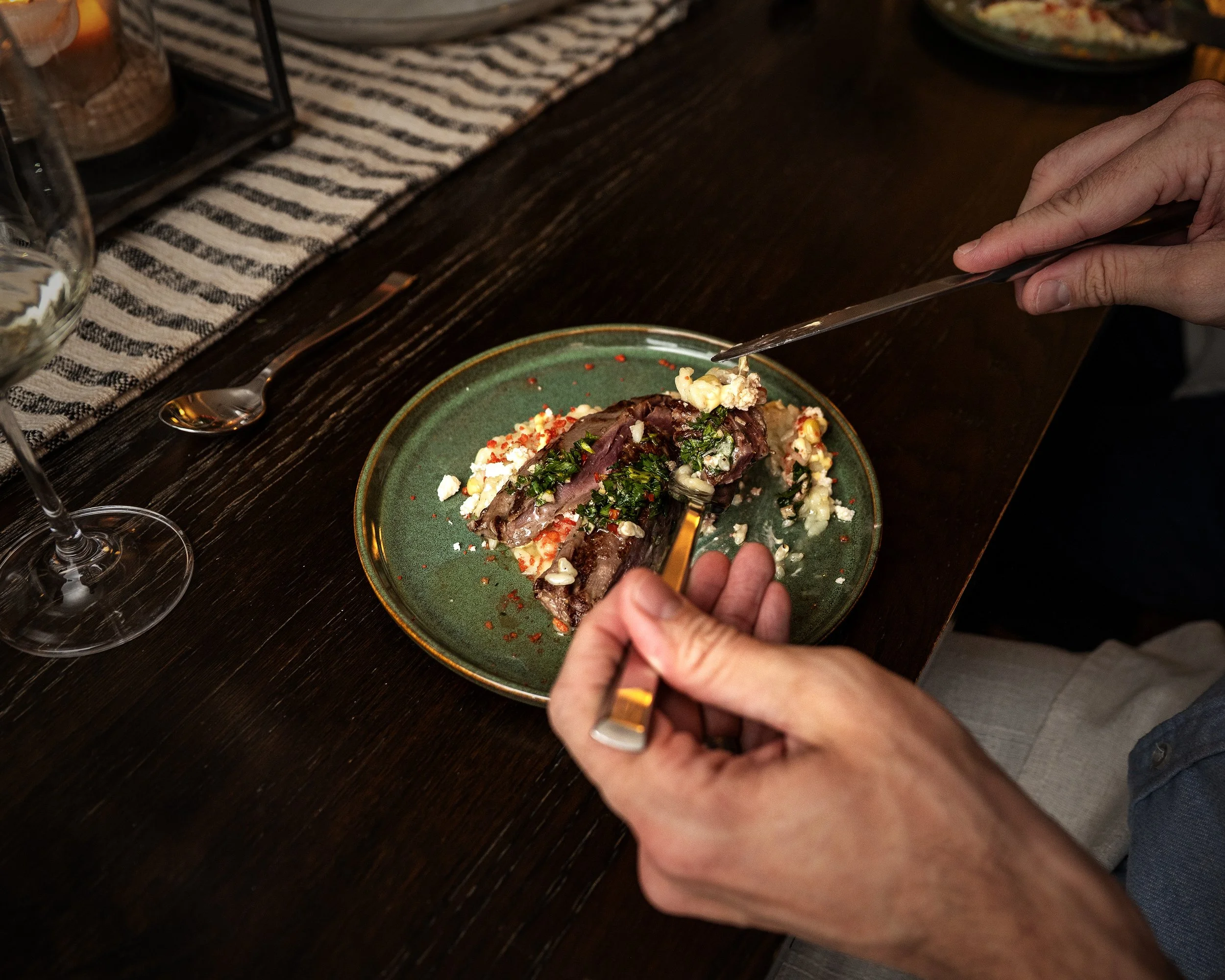 Person using tongs to serve food from a green plate containing meat, vegetables, and garnishes on a dark wooden table with a striped cloth and wine glass nearby.