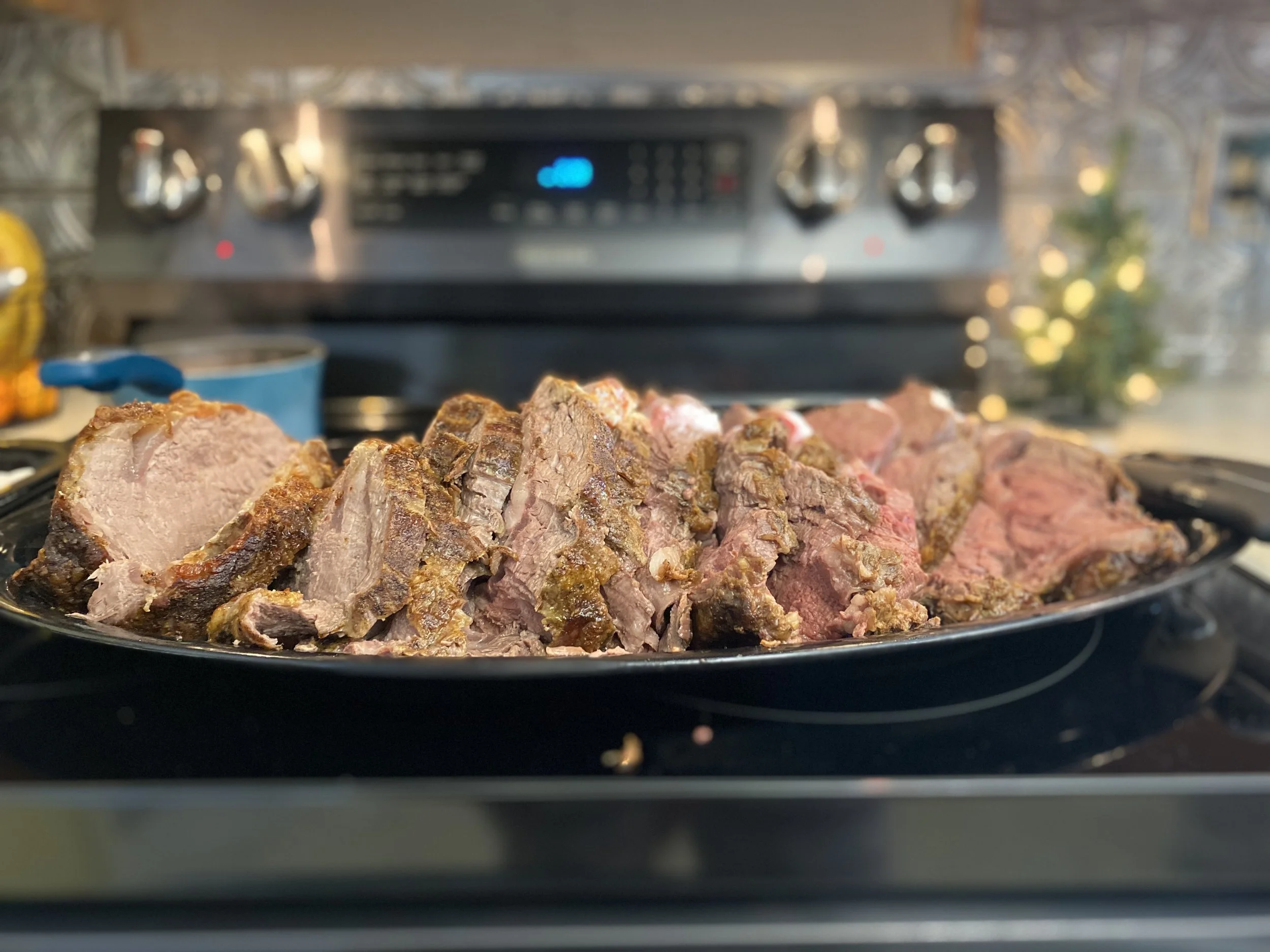 Close-up of cooked meat, including beef and pork, resting on a black tray, with a kitchen stove in the background.