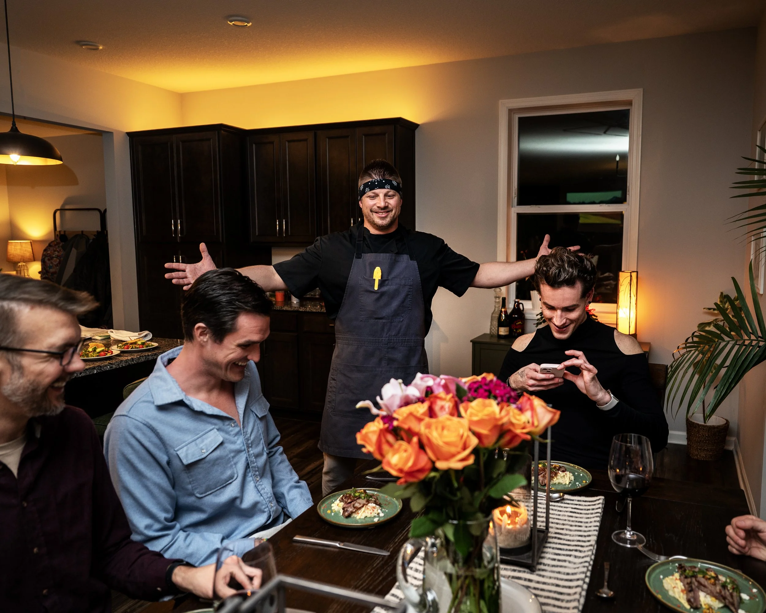 A man wearing a black bandana and apron stands with his arms open, smiling during a dinner party. Several guests are seated at a dining table with plates of food, wine glasses, and a floral centerpiece. The room is warmly lit, with dark cabinets and 