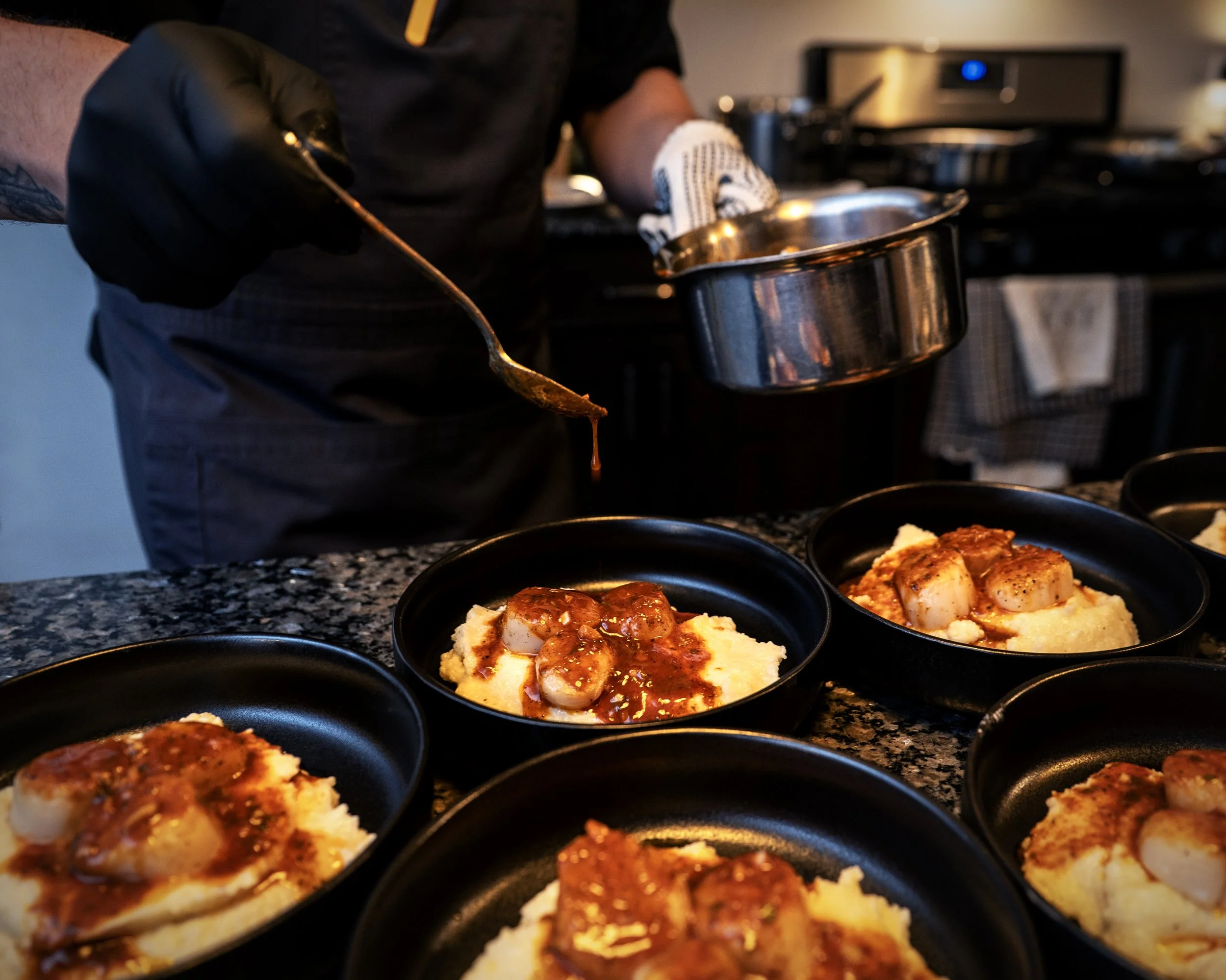 A chef wearing black gloves is preparing small dishes of food, garnished with sauce, in a professional kitchen. The dishes are placed on a granite countertop.