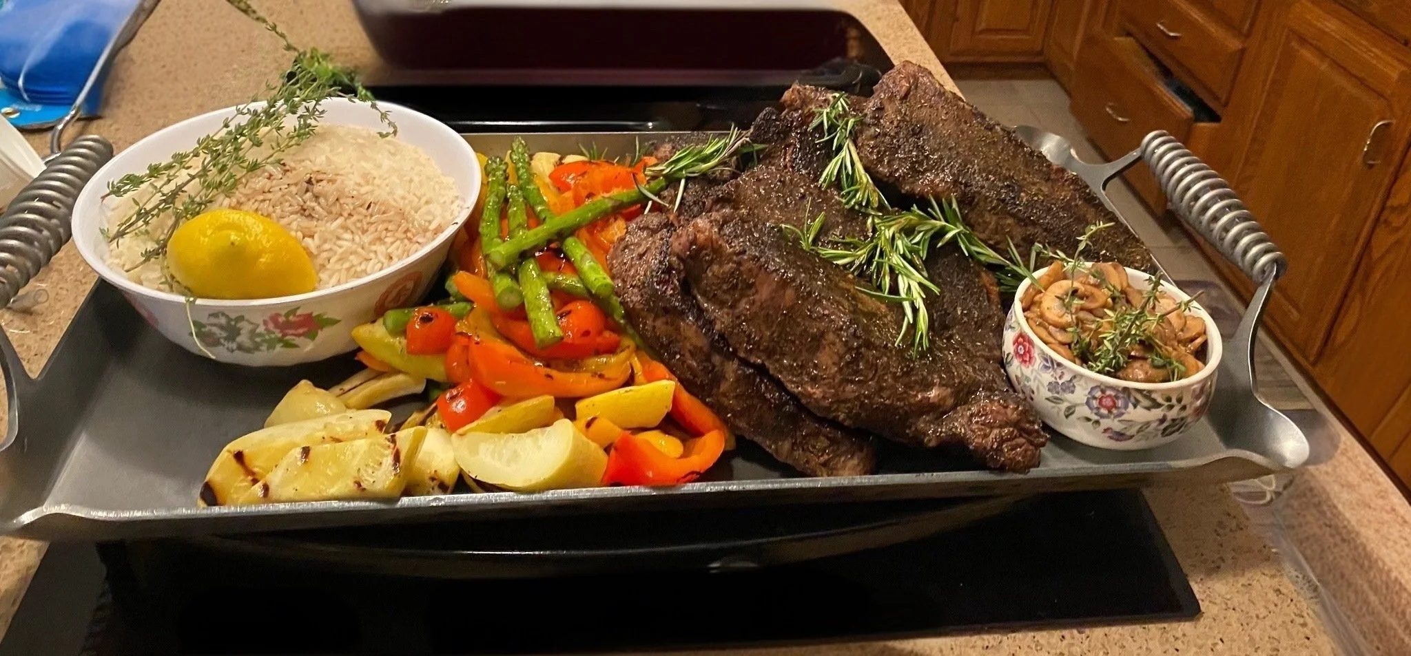 A tray with cooked ribs garnished with rosemary, a bowl of rice with a lemon wedge and sprigs of herbs, roasted vegetables including yellow and red peppers and zucchini, roasted potatoes, and a small bowl of cooked mushrooms garnished with herbs.