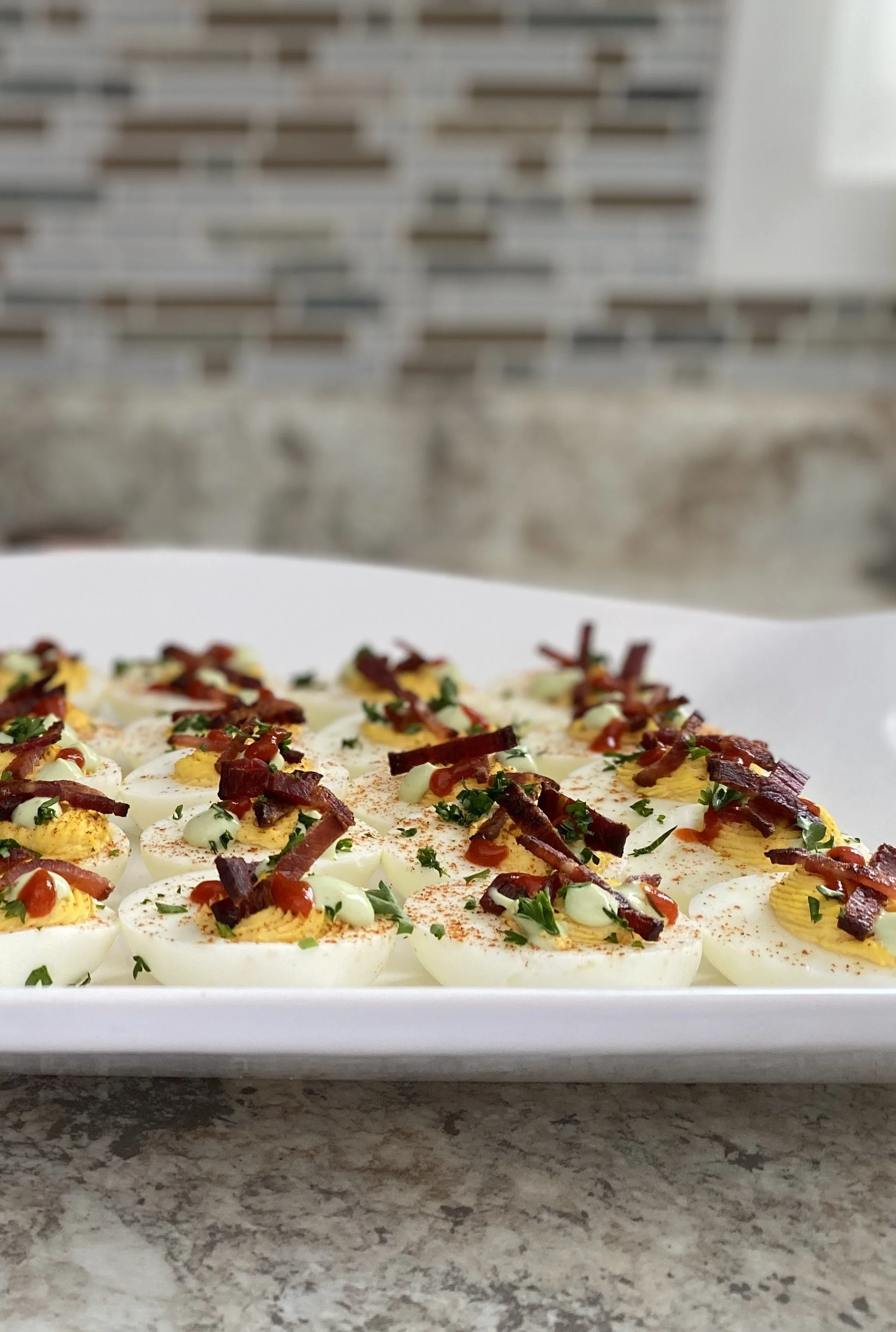 A tray of sliced hard-boiled eggs topped with bacon bits, parsley, and drizzled with sauces, with a brick wall in the background.