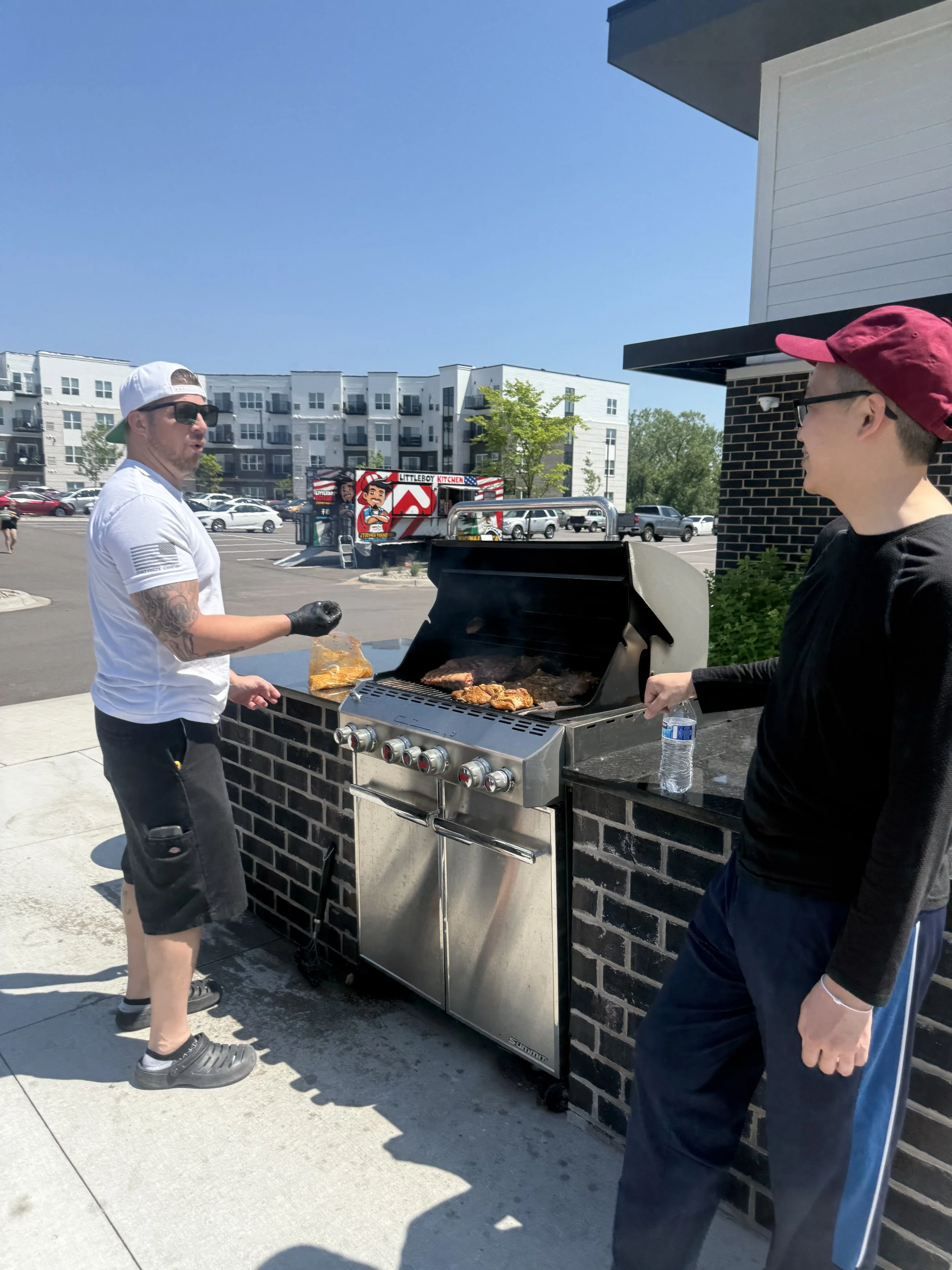 Two men are standing outdoors, grilling meat on a barbecue grill. One man is wearing a white cap backwards, sunglasses, a white T-shirt, and shorts. The other man is wearing a red cap sideways, glasses, and a black shirt. There is a parking lot with 