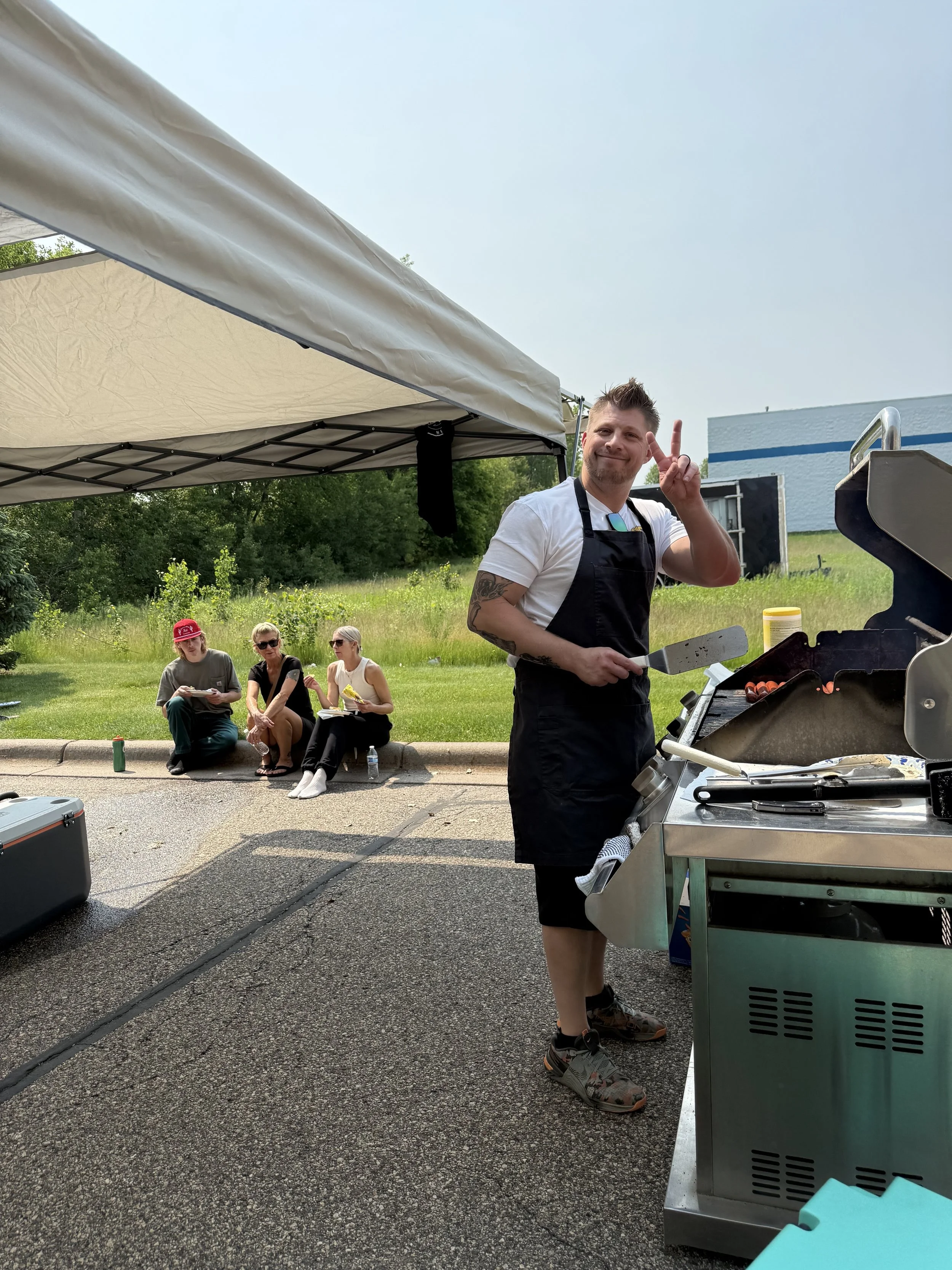 Man grilling at an outdoor event with a barbecue grill, while three women sit on the ground nearby under a tent, with trees and a building in the background.