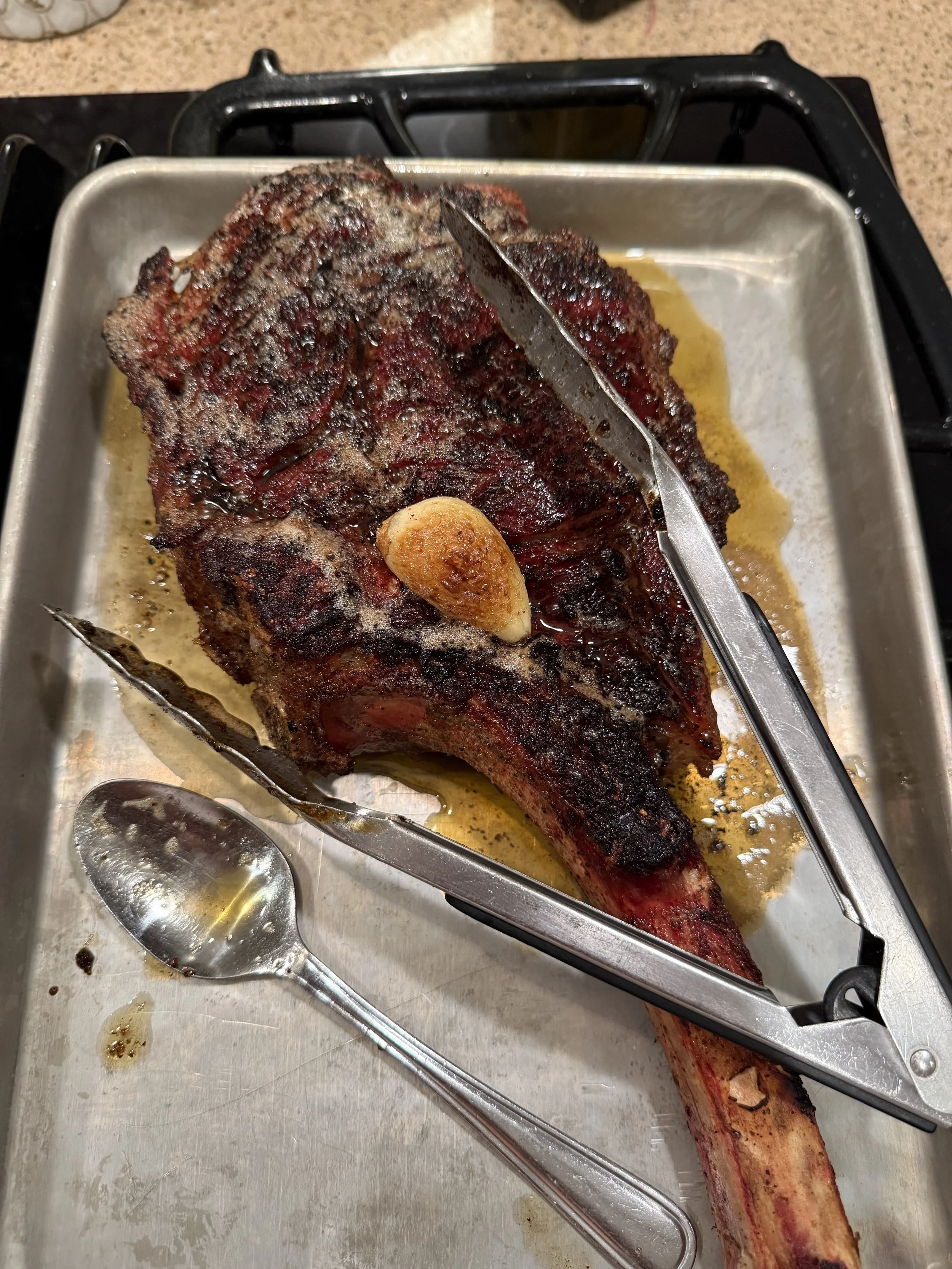 A large, cooked T-bone steak with a visible bone, resting in a metal pan, with a knife inserted into it and a garlic clove on top. There are serving utensils on the pan and some juices surrounding the steak.