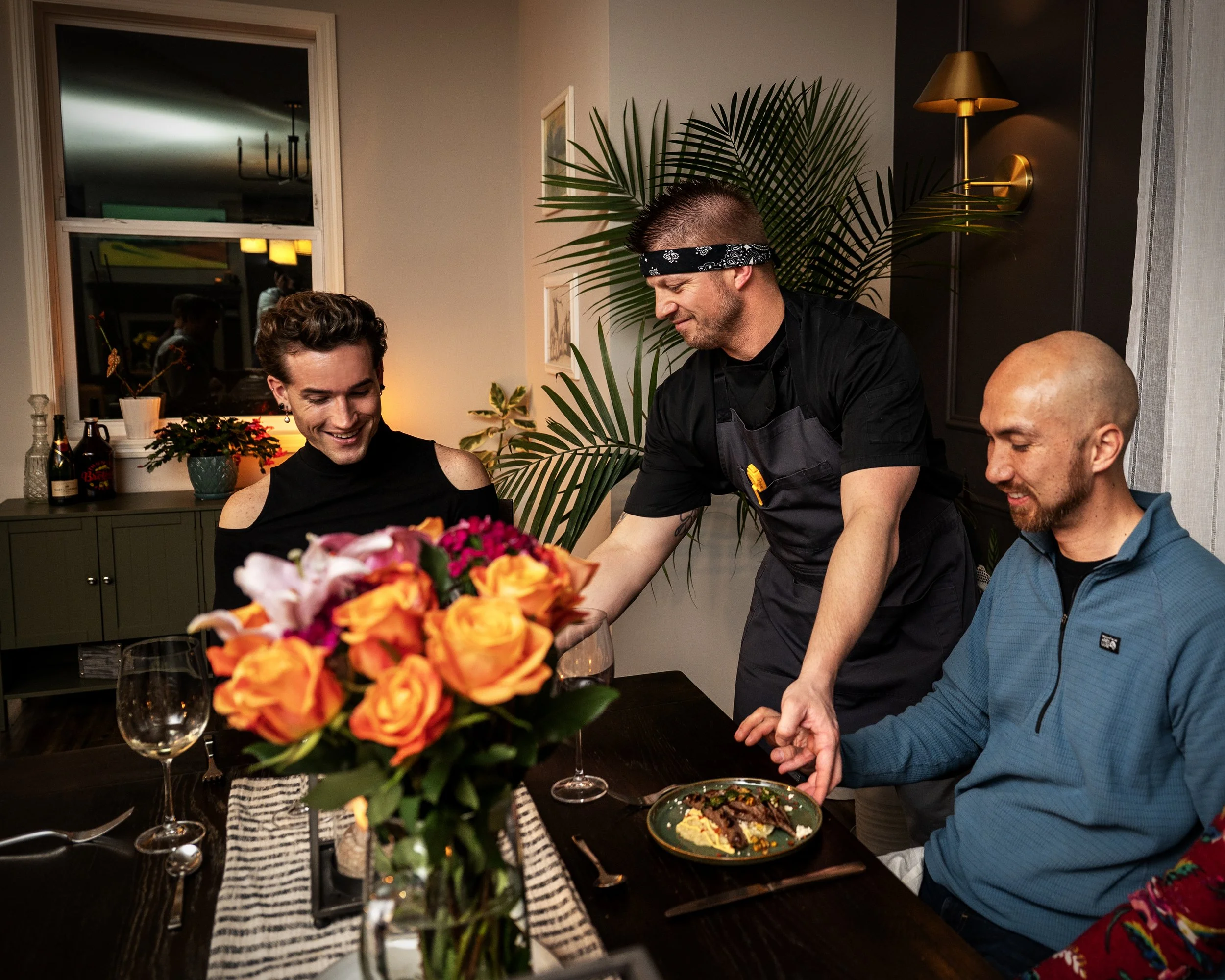People gather around a dining table with a centerpiece of orange and pink flowers, enjoying a meal in a warmly lit room with green cabinets, potted plants, and wall art.
