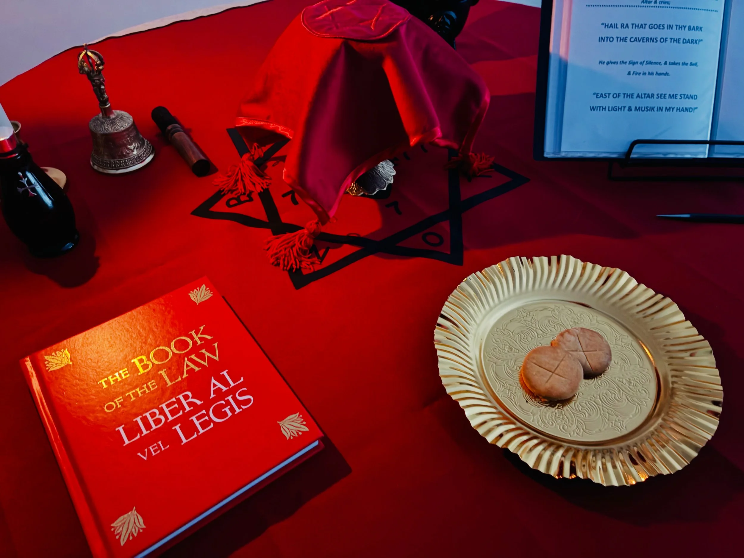 A red table with a book titled 'The Book of the Law of the LiberALS' by Joel LLEGS, a brass bell, a black vial, a black pen, a folded red cloth with black tassels, a framed sheet with blue text, and a paper plate with three cookies.