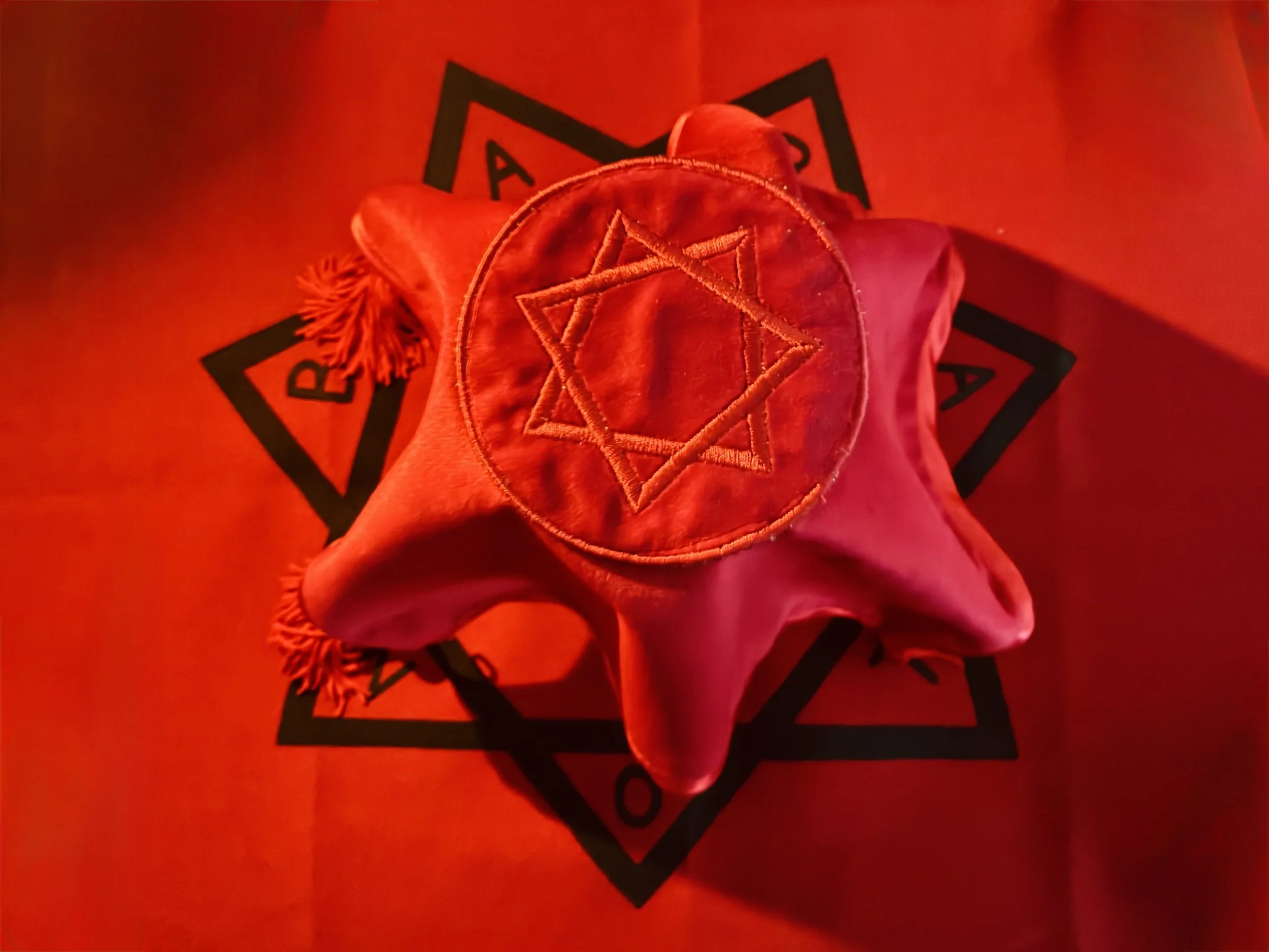 A piece of cloth with embroidered pentagrams and designs, illuminated in red light, placed on a surface with a black and red symbol.
