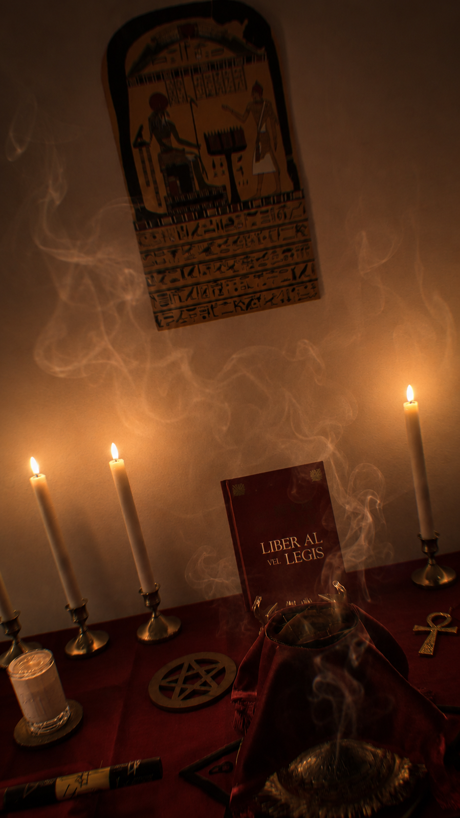 A table with lit candles, incense smoke, a pentagram, a candle in a glass holder, a red cloth, a black marker, and a book titled 'Liber Al Vel Legis'. On the wall is an Egyptian-style painting of two figures and hieroglyphs.