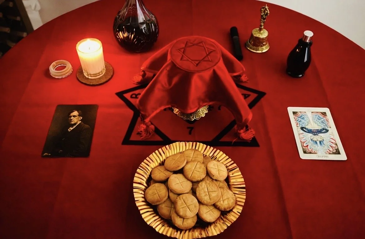 A table with a red cloth, a plate of cookies, tarot or oracle cards, a candle, a black bottle, a small golden chalice and a red velvet cover with an embroidered star.