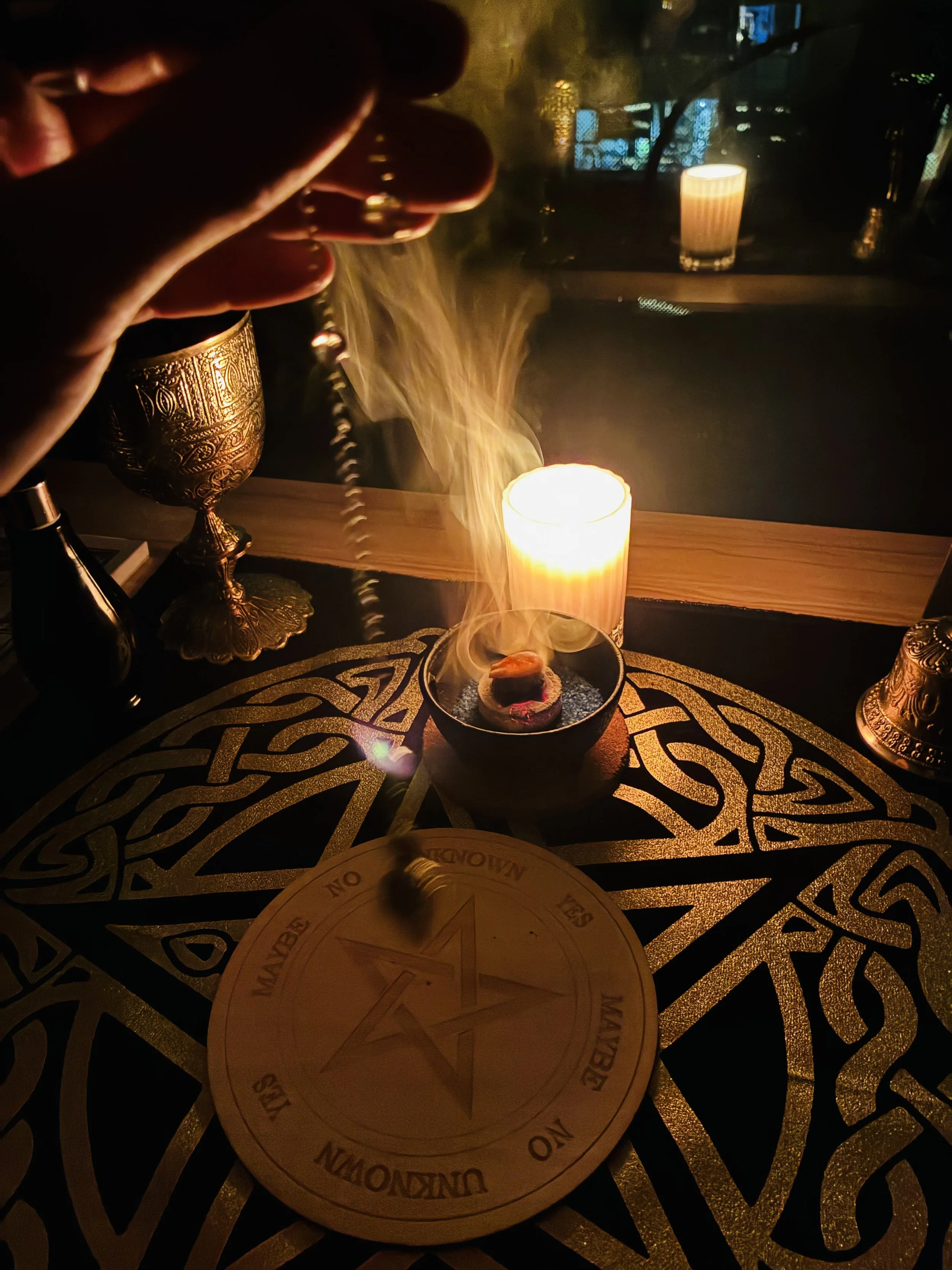 A person holding a telephone receiver at a table decorated with candles, a brass ornamental vase, and a round wooden object with a pentagram and the words 'The Bewitching of the Unknow' around its edge. The setting is dimly lit with warm candlelight and has a mystical, occult atmosphere.