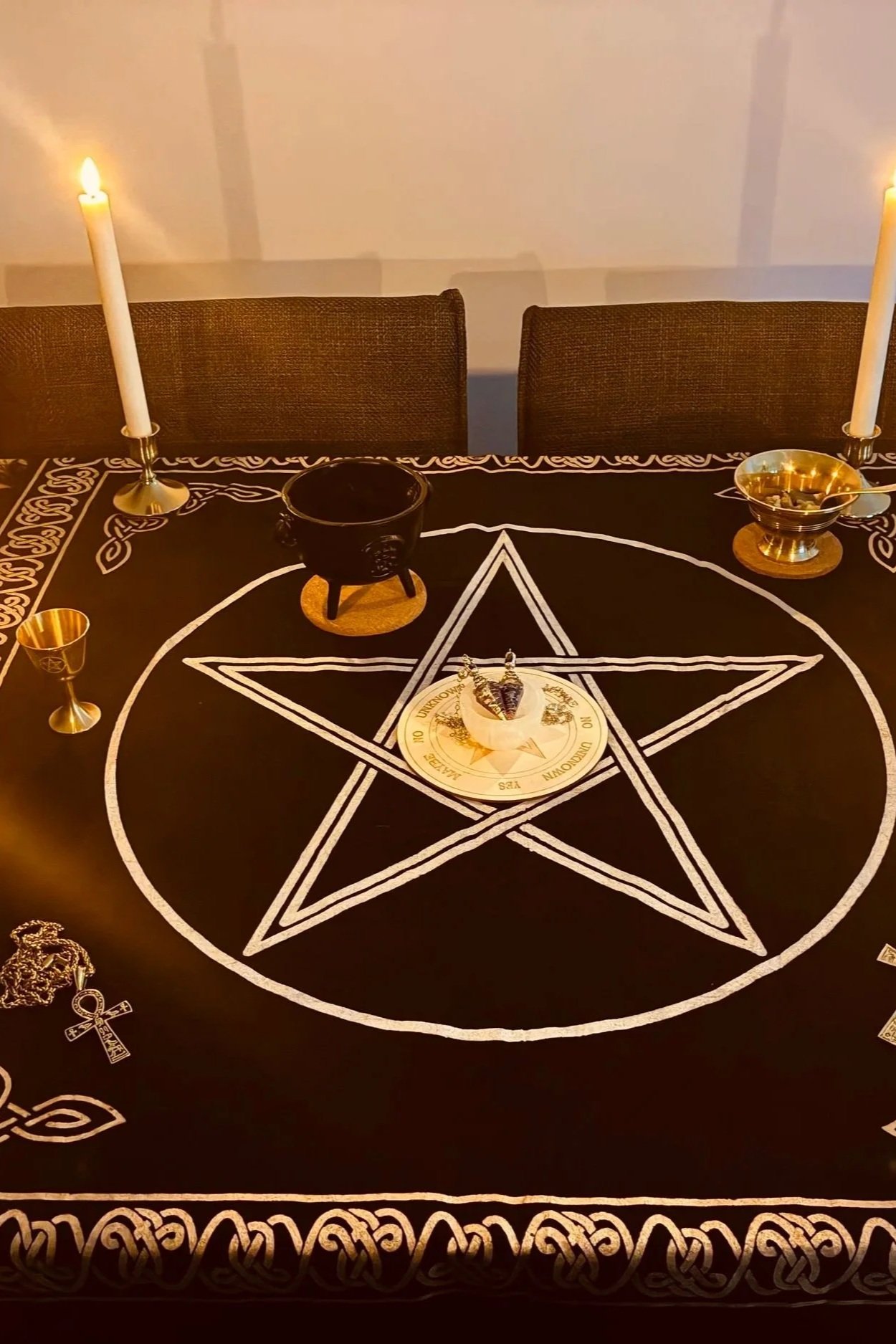 Table covered with a black cloth featuring white occult symbols, including a pentagram in the center and other mystical designs. The table has two lit candles, a black cauldron, a gold bowl with a spoon, and a gold goblet, all arranged in a ritualistic setting.