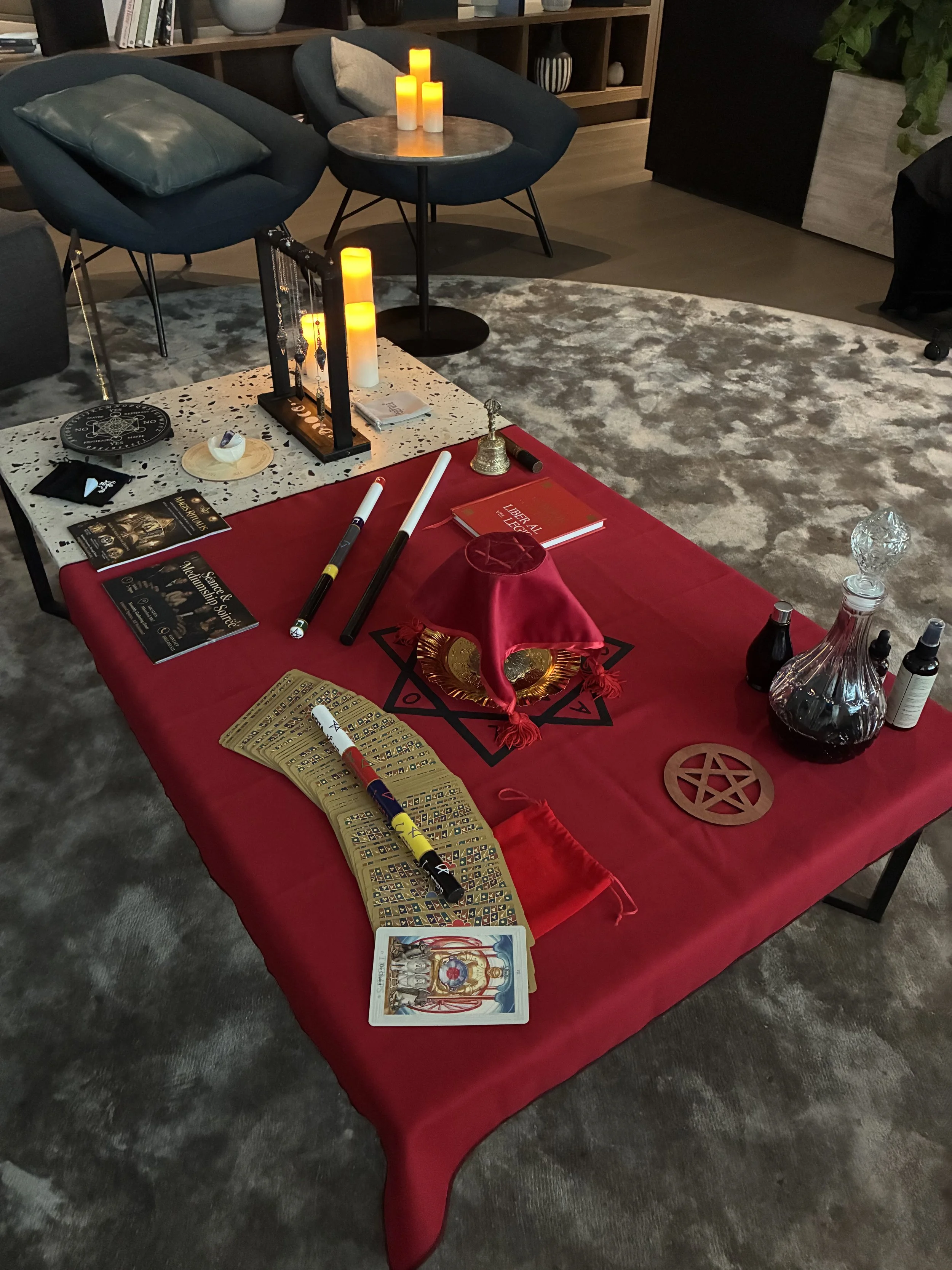 A table covered with a red cloth holds tarot and oracle cards, a crystal ball, and a red velvet bag. A pentagram symbol is drawn on the cloth, and a magic staff or wand lies across the table. In the background, there are two blue armchairs, a small side table with candles, and shelves with various decorative items.