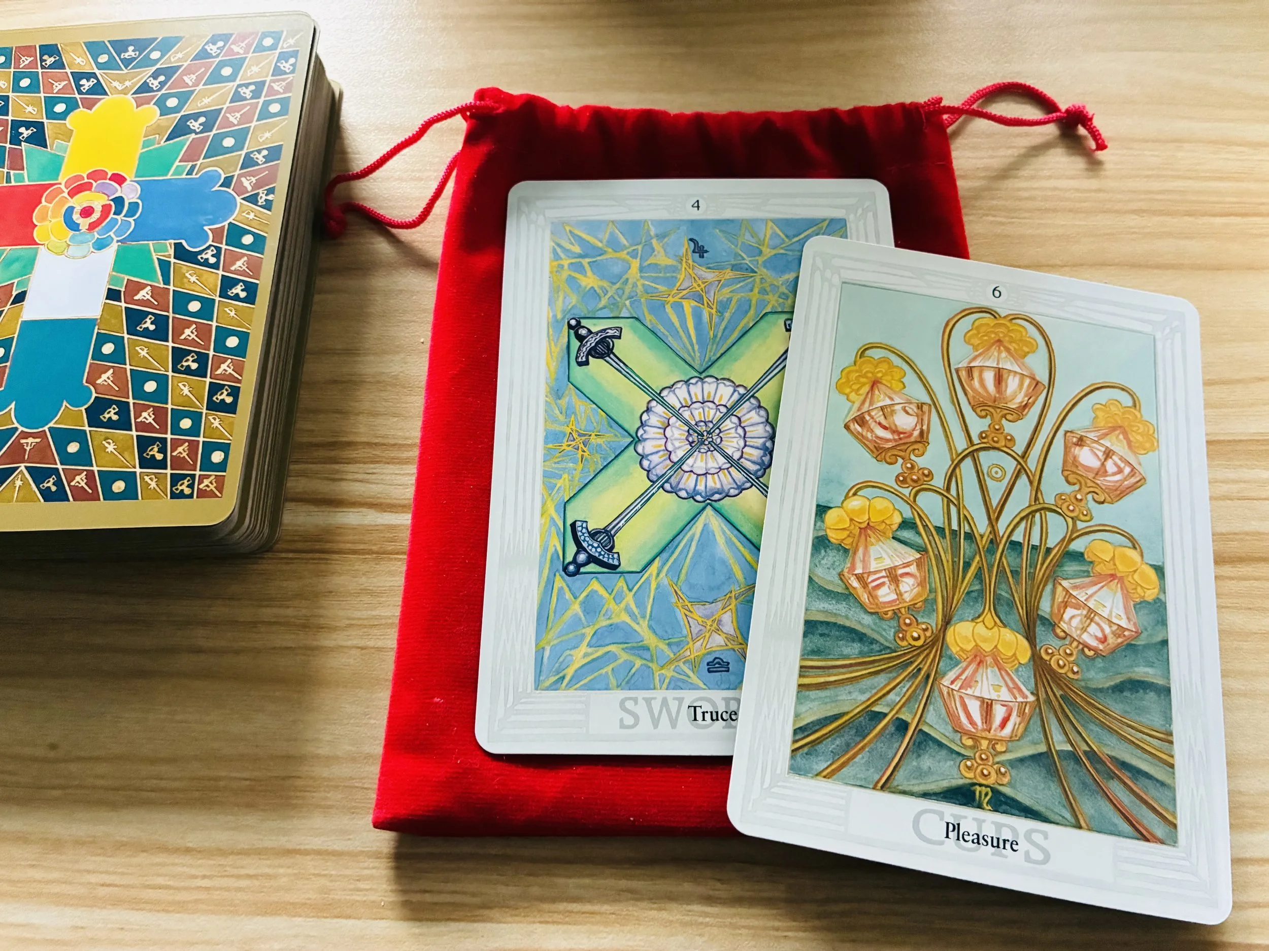 A deck of cards with colorful illustrations, two cards face up showing the Two of Swords and the Three of Cups, placed on a red velvet pouch on a wooden surface.