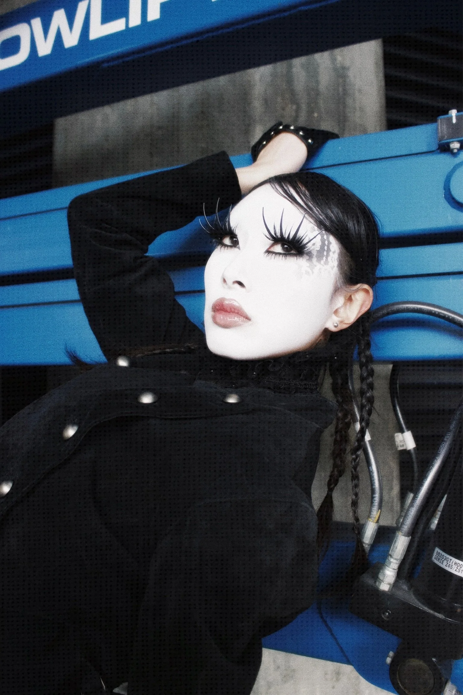 A woman with dramatic makeup and long lashes leaning against a blue bowling alley lane, resting her hand on the surface, with braided hair and wearing a black outfit with silver studs.
