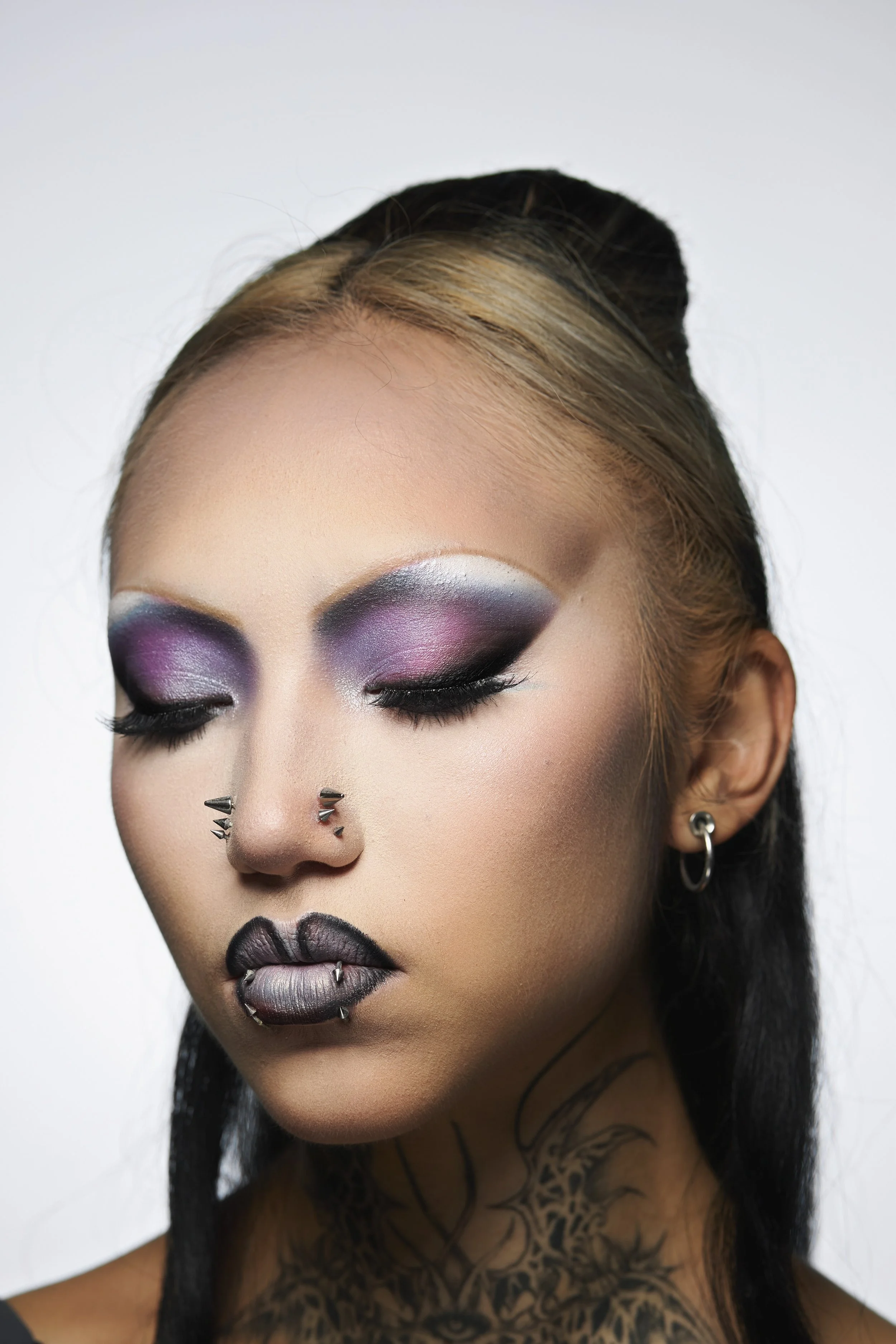 Close-up portrait of a woman with bold makeup, tattoos, and piercings, with her eyes closed.
