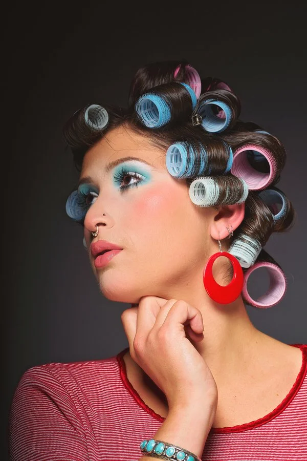 A woman with hair rollers, vibrant blue eyeshadow, pink lipstick, a red earring, and wearing a red and white striped top.