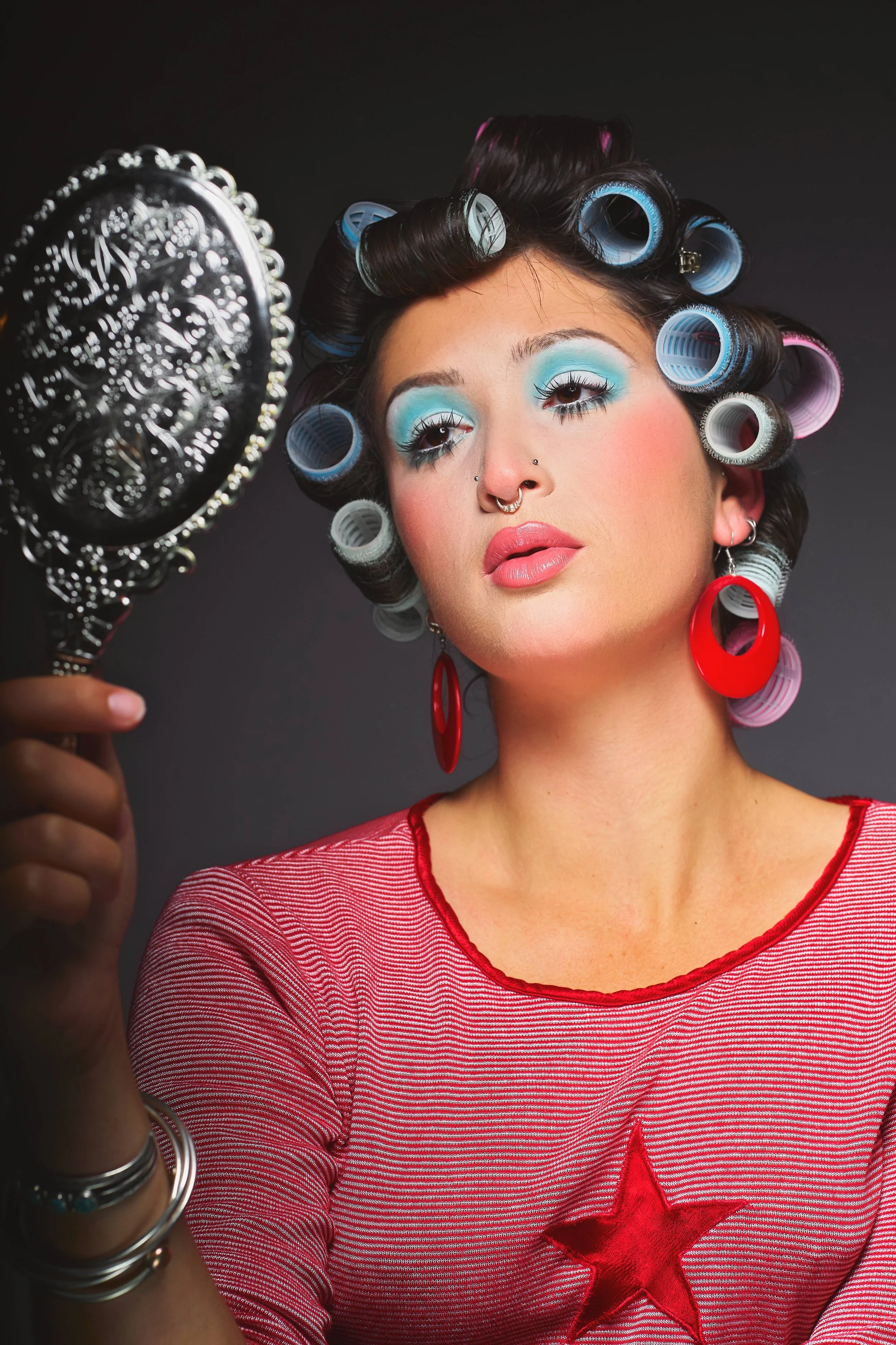 Woman with curled hair in rollers applying makeup, wearing bright makeup, red star shirt, large red earrings, looking in mirror.