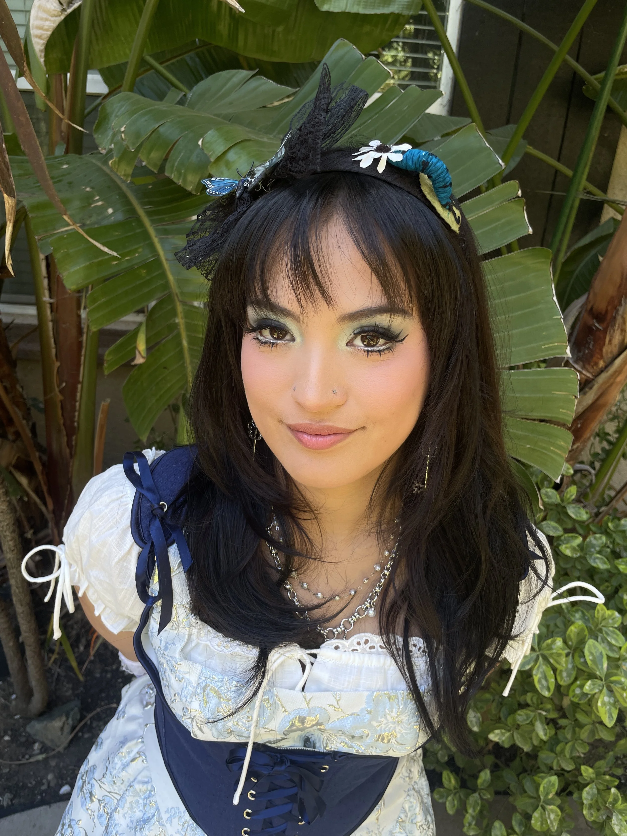 A woman with black hair, makeup, and jewelry, standing outdoors in front of green tropical plants, wearing a white embroidered dress with blue and gold details, and a headband with a butterfly and flower embellishments.