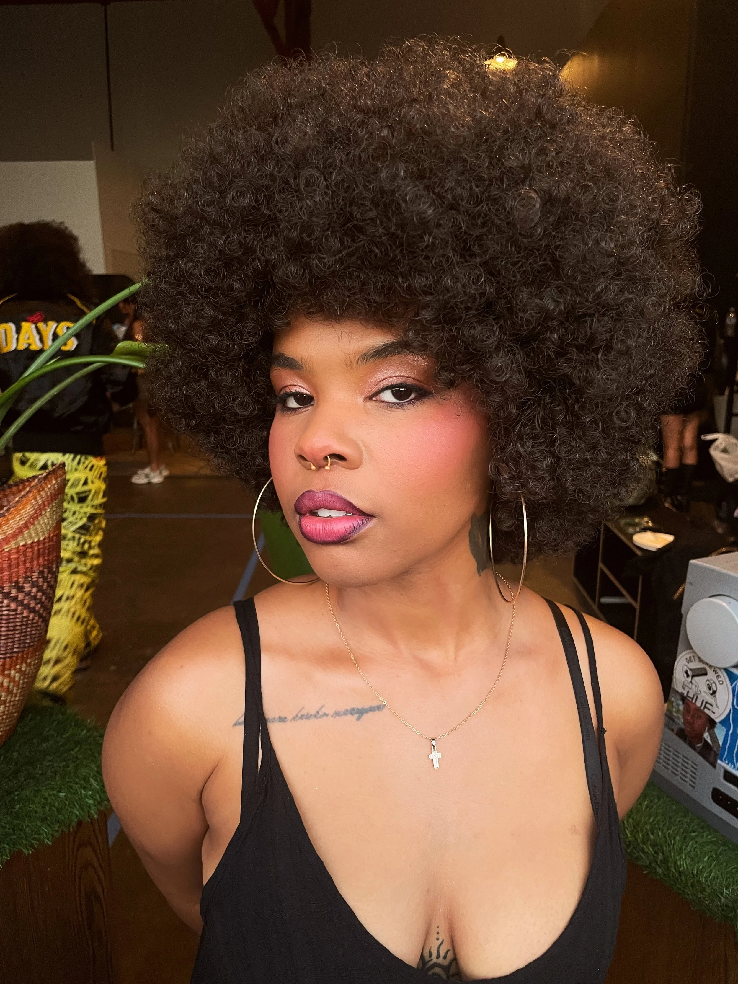 A woman with large, voluminous curly hair, wearing a black spaghetti strap top, hoop earrings, a cross necklace, and makeup with pink lipstick, posing indoors.