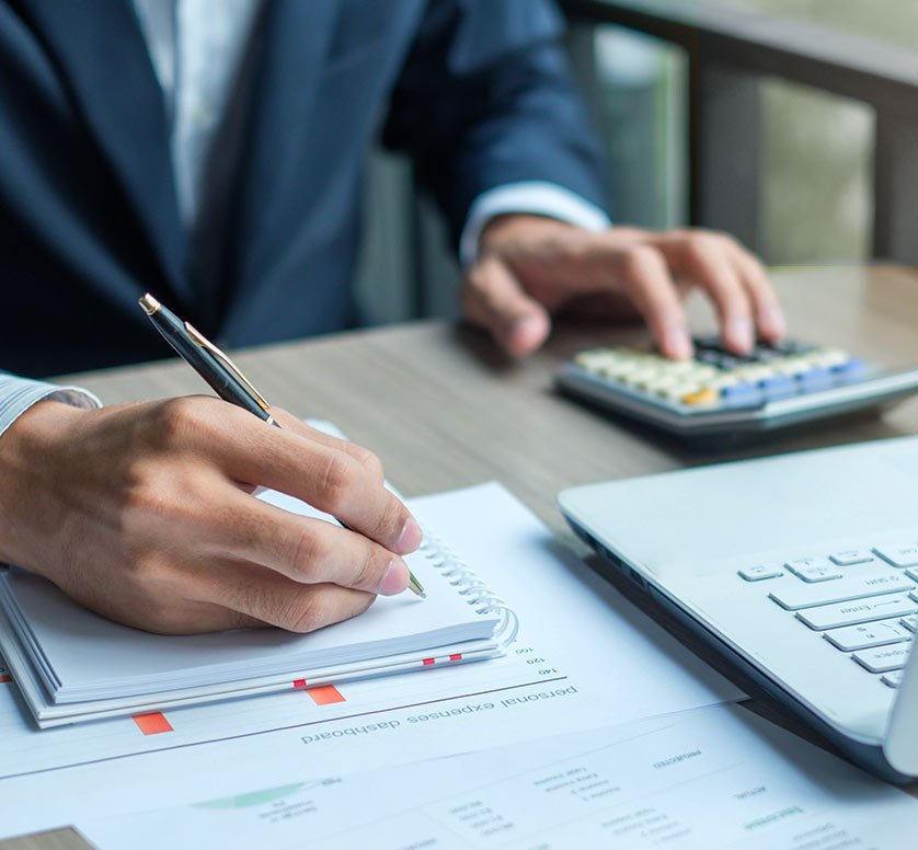 Person in a suit writing in a notebook at a desk with a calculator, laptop, and financial documents.
