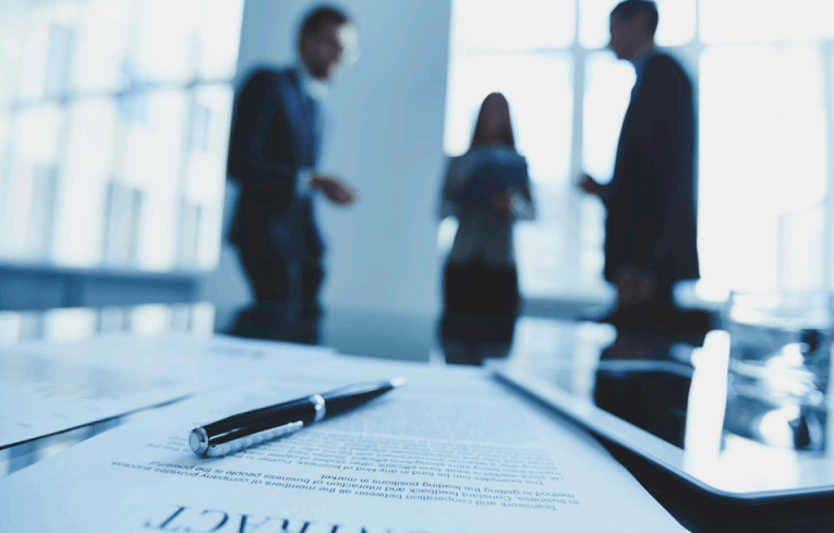 Blurred image of three business professionals standing and talking in a bright office space with large windows, with documents and a pen on the table in the foreground.