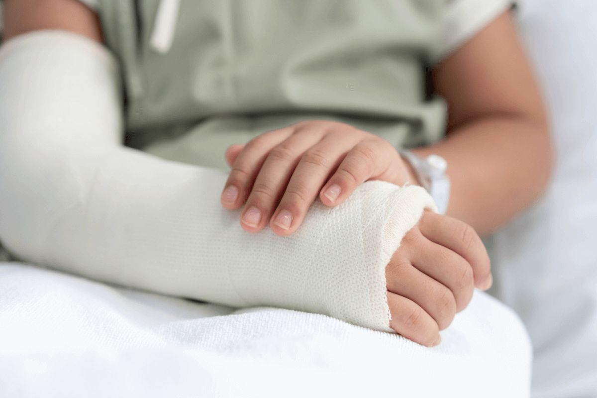 A child's hand with a cast on their arm, being gently held by an adult's hand, in a medical setting.