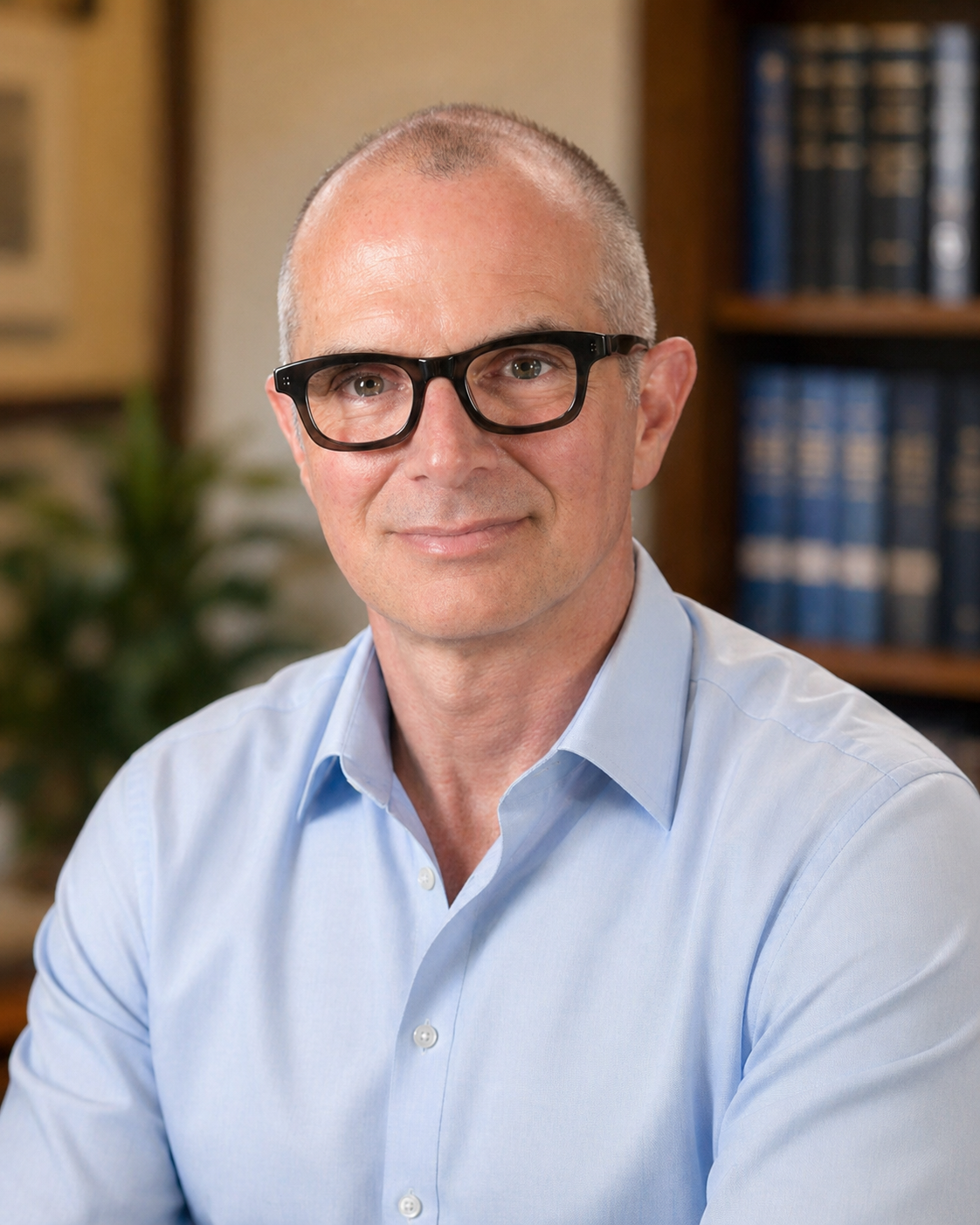 A middle-aged man with a shaved head and black glasses smiling at the camera. He is wearing a light blue dress shirt and is seated indoors with a background of bookshelves and a blurred plant.