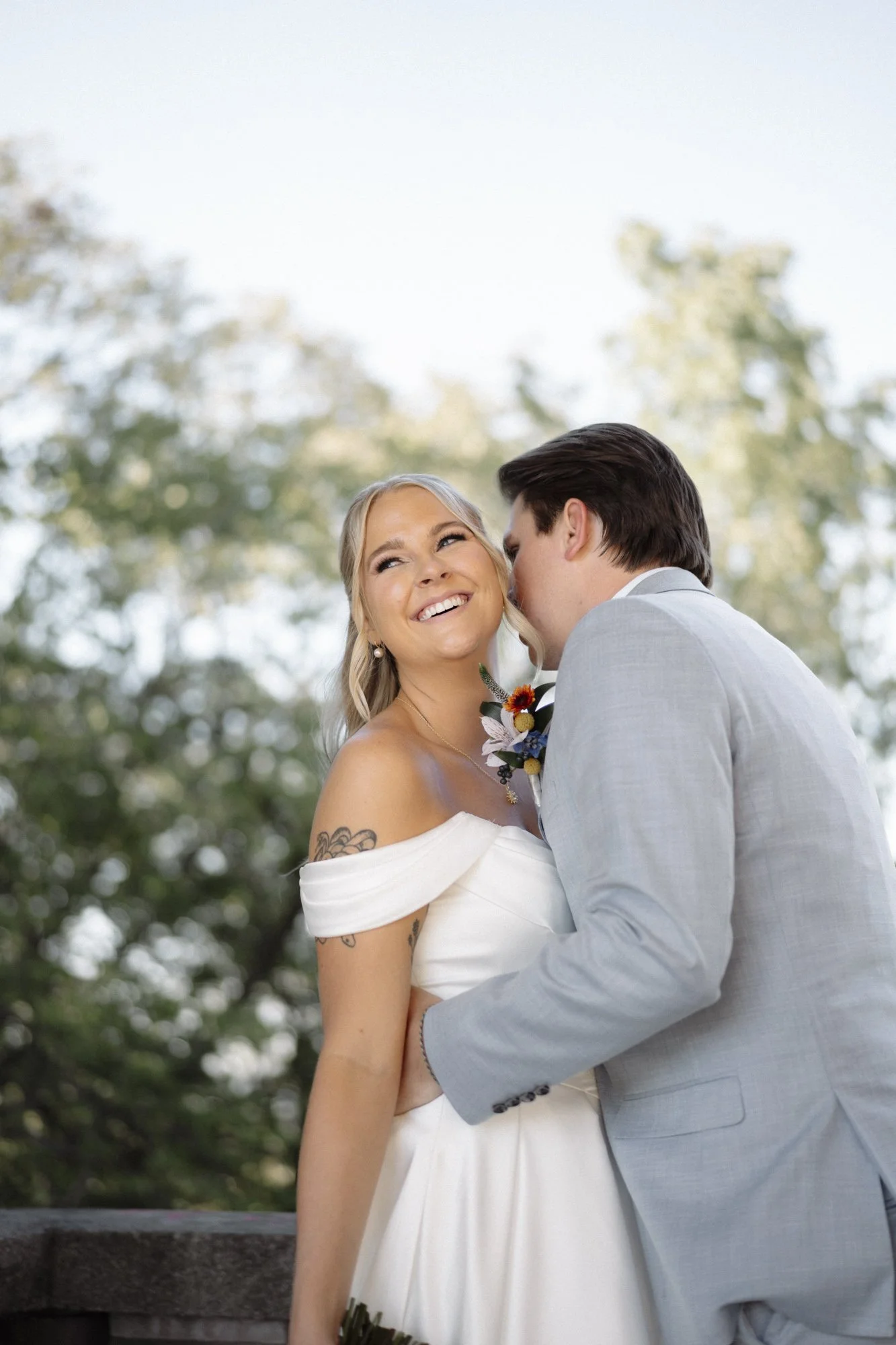 A bride and groom kissing outdoors, with the bride's smiling face visible and wearing a white off-shoulder wedding dress, while the groom wears a light gray suit.