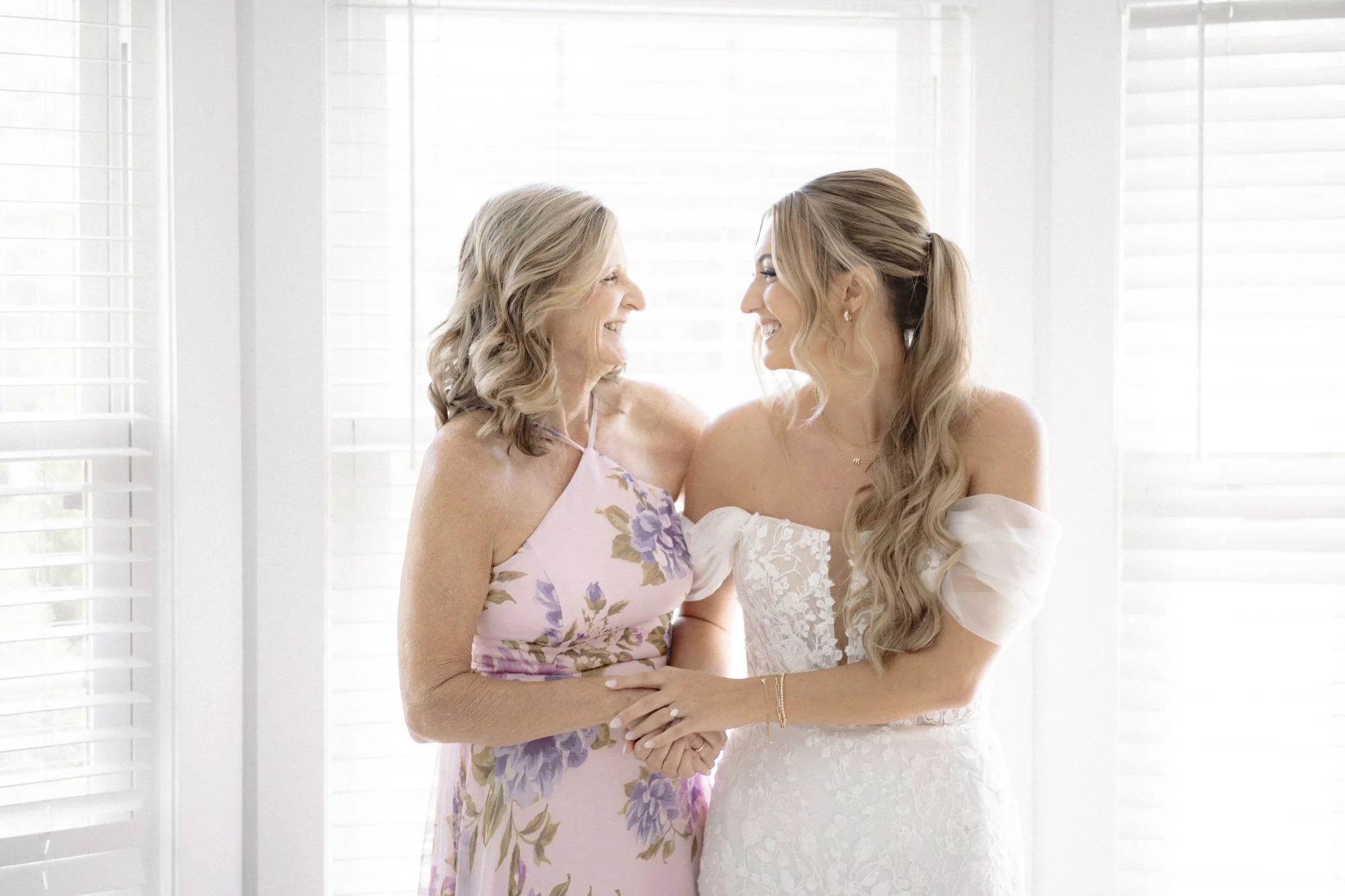 A young woman and an elderly woman smiling and holding hands in front of white blinds, with sunlight filtering through.