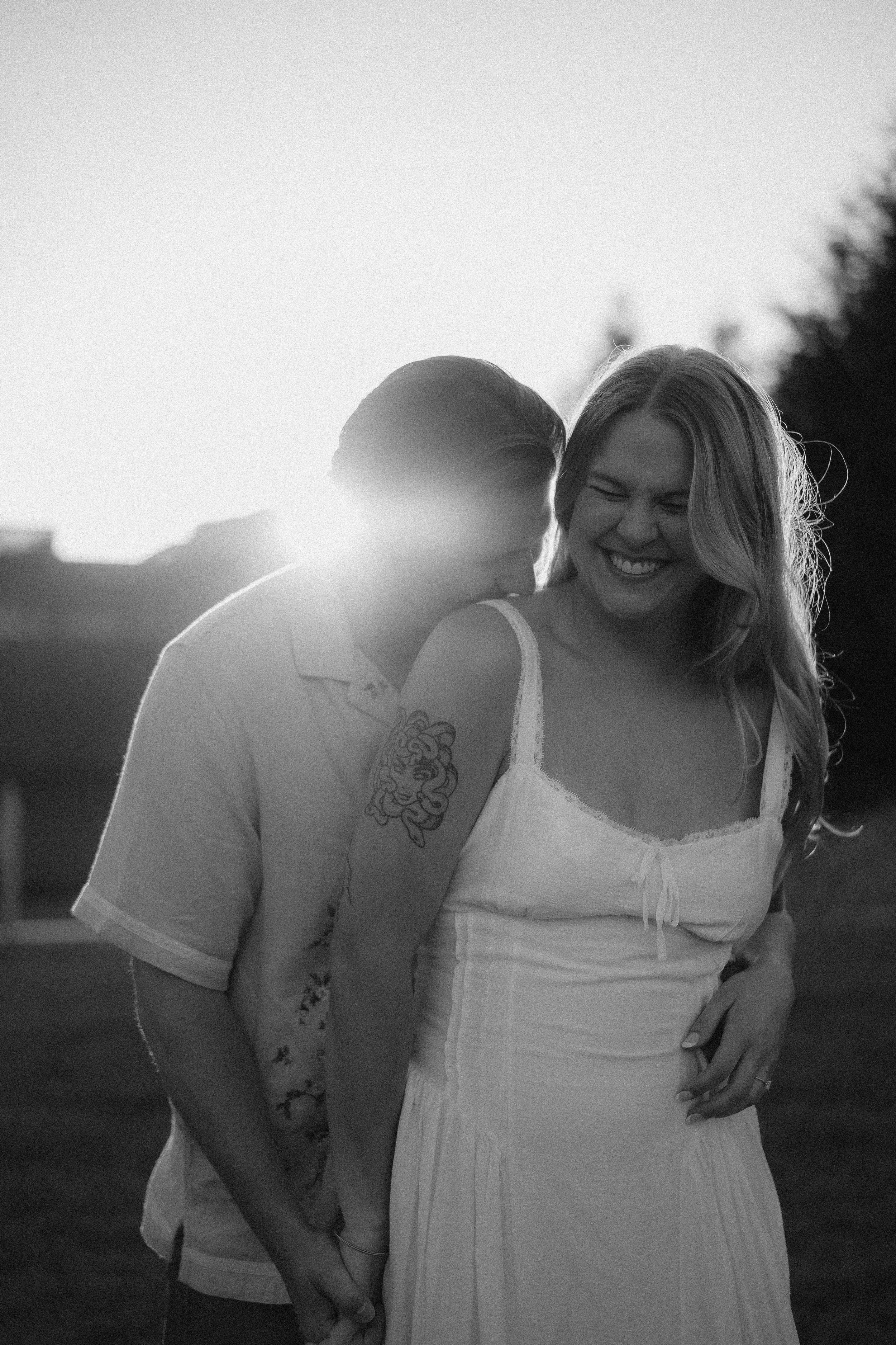 A woman with long hair in a white dress smiling and laughing while a man in a short-sleeved shirt leans in close to her, outdoors in sunlight.