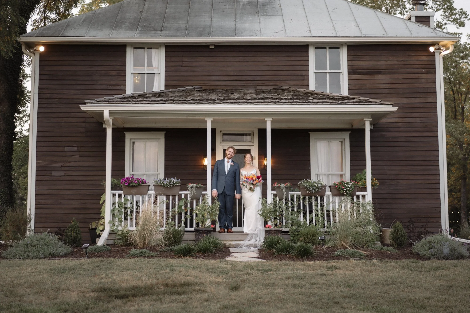 A bride and groom standing on a porch of a dark brown, two-story house with white trim. The bride is in a white wedding dress holding a colorful bouquet, and the groom is in a dark suit with a bow tie. Flowers and plants are in front of the porch, and the house has a metal roof and lit wall-mounted lights beside the door. It appears to be around dusk or evening.