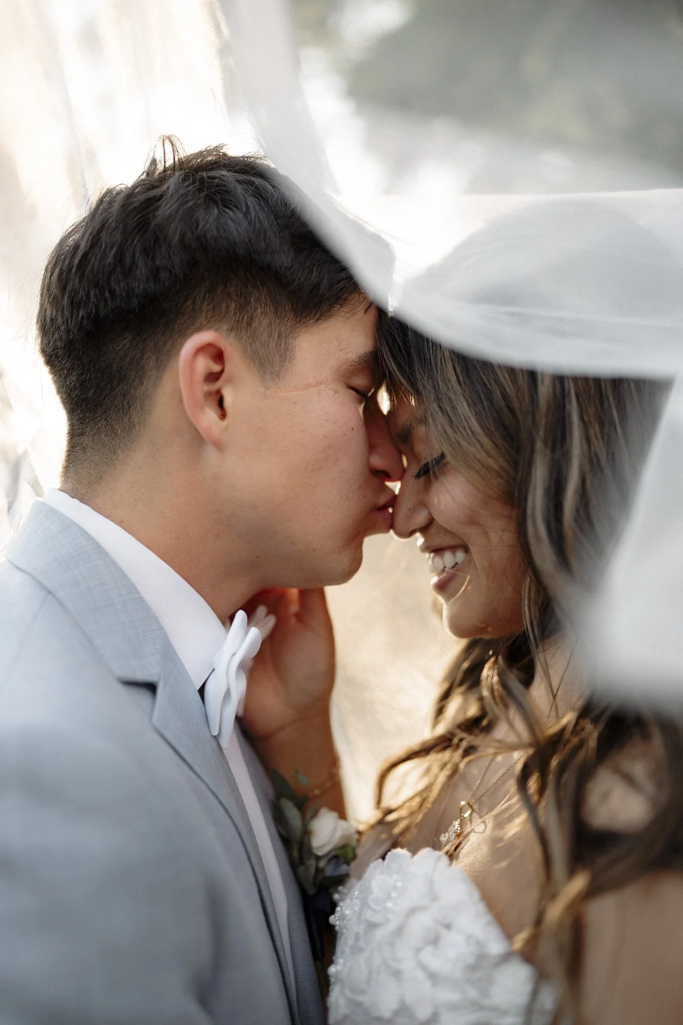 A couple shares a close, intimate moment on their wedding day, with foreheads touching and eyes closed, under a white veil, smiling and dressed in wedding attire.