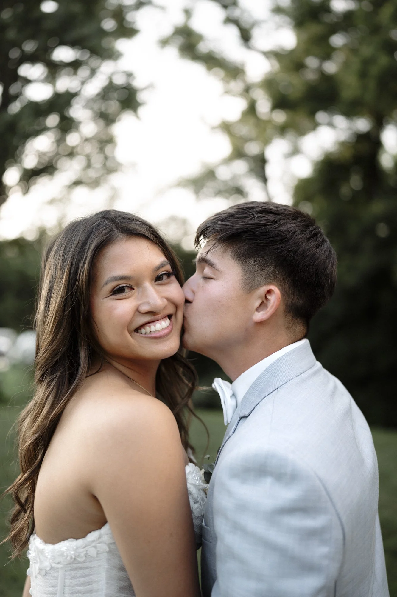 A young woman in a white wedding dress smiling while a young man in a light gray tuxedo kisses her on the cheek outdoors with trees and sky in the background.