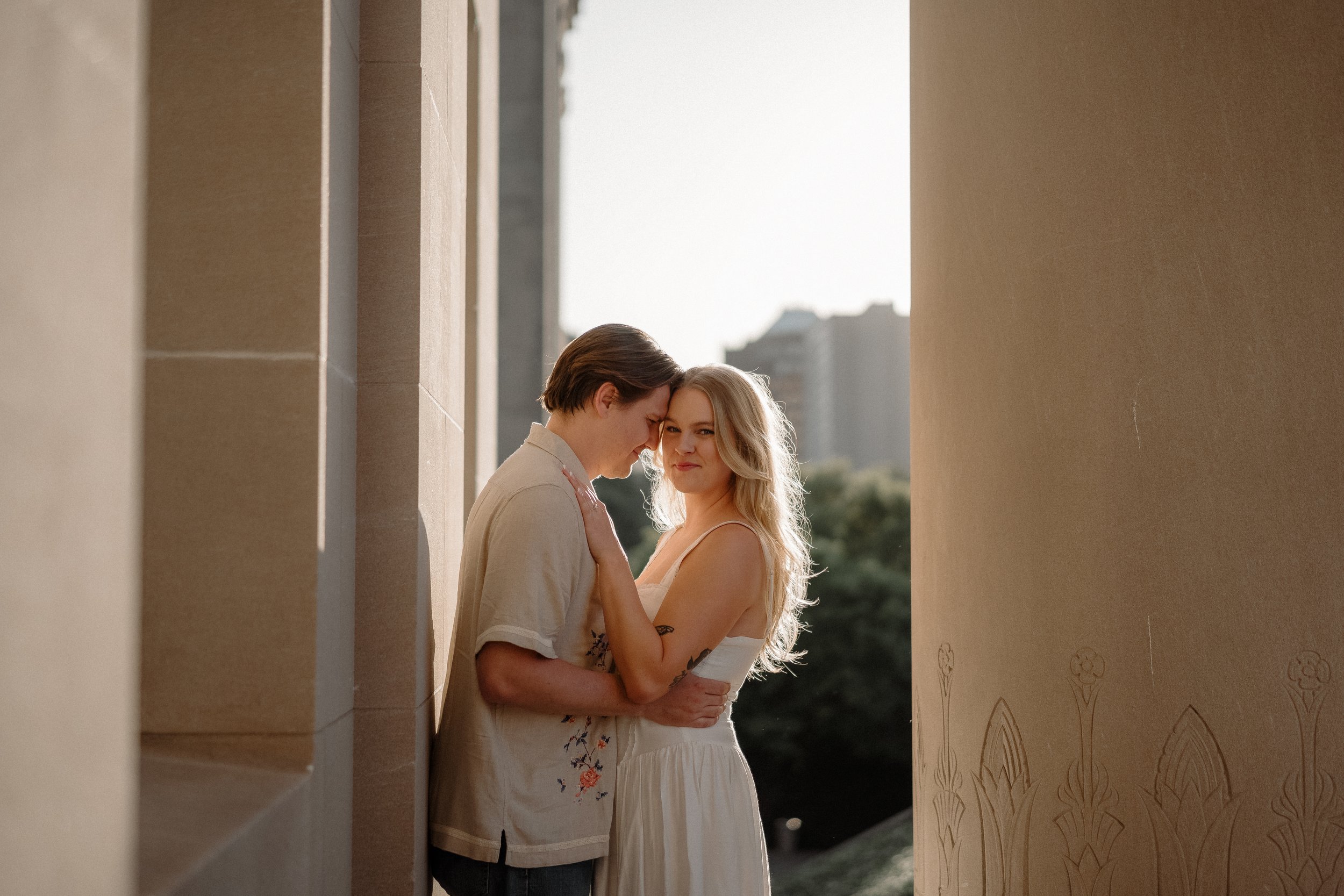 A couple sharing an intimate moment, with foreheads touching and eyes closed, standing close in a narrow passageway between two buildings during sunset.