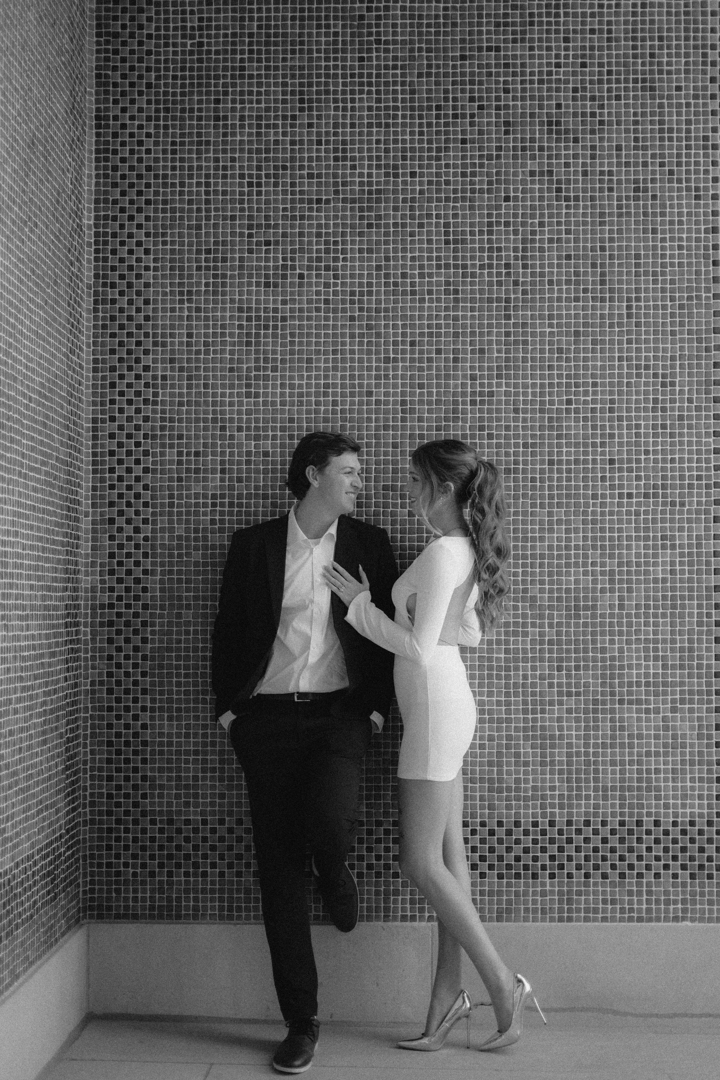 A black-and-white photo of a man and woman standing close together against a tiled wall, smiling and looking at each other. The man is wearing a suit with his hands in his pockets, and the woman is in a fitted dress and high heels, touching the man's chest.