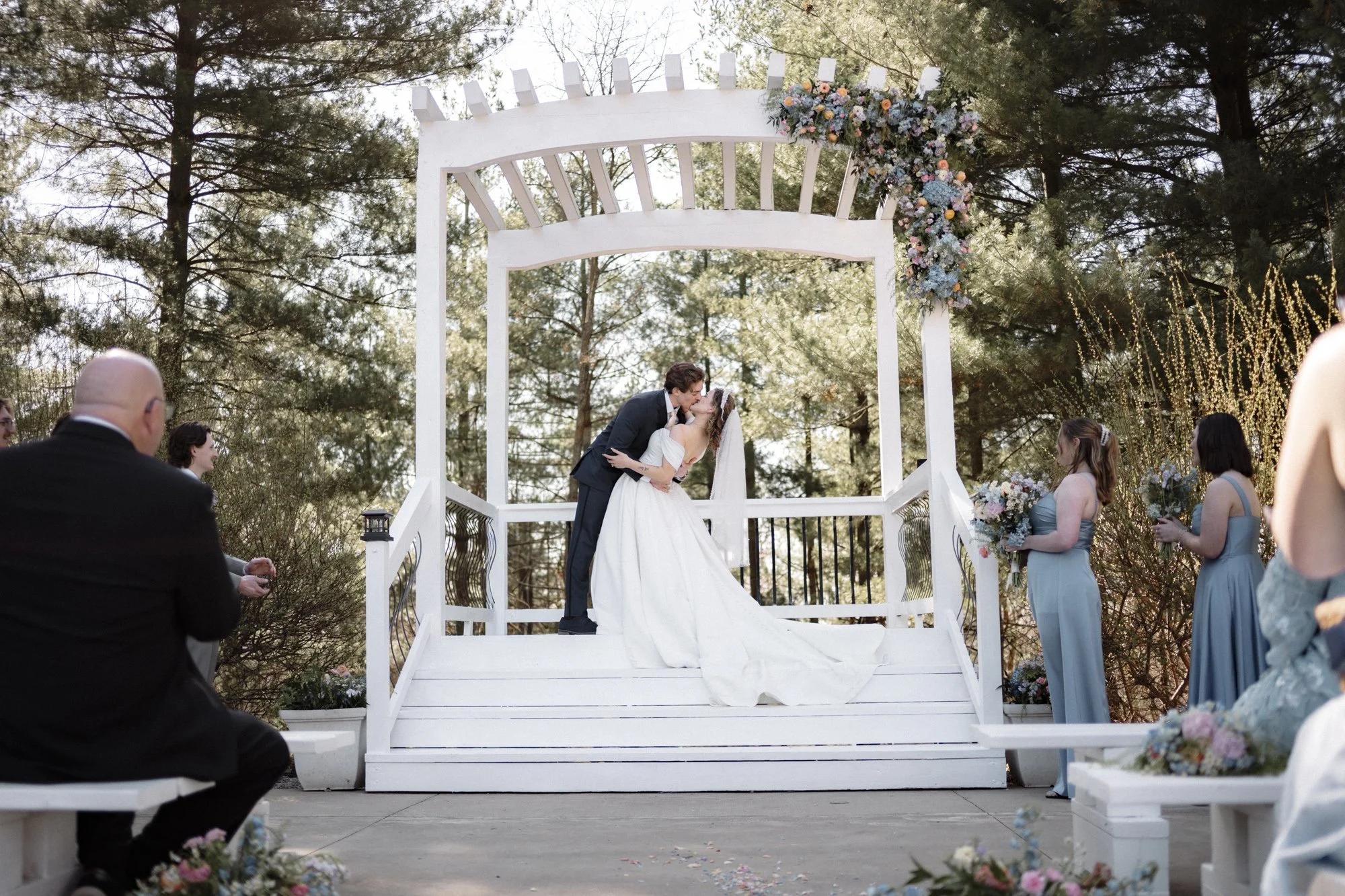 A bride and groom share a kiss on a white gazebo decorated with flowers at an outdoor wedding ceremony in a wooded area.