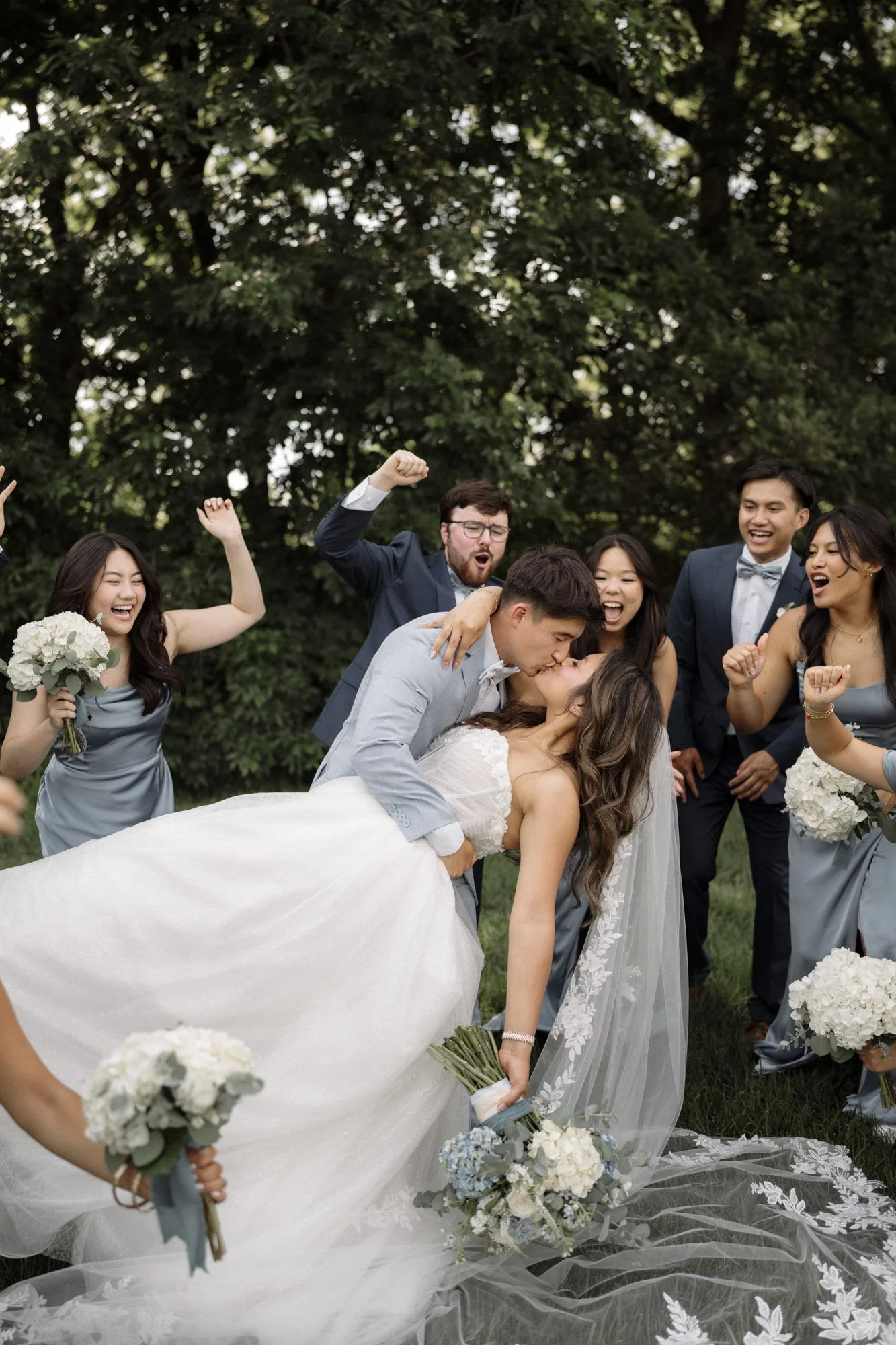 A wedding group photo with the bride and groom kissing, surrounded by friends celebrating outdoors during daytime.