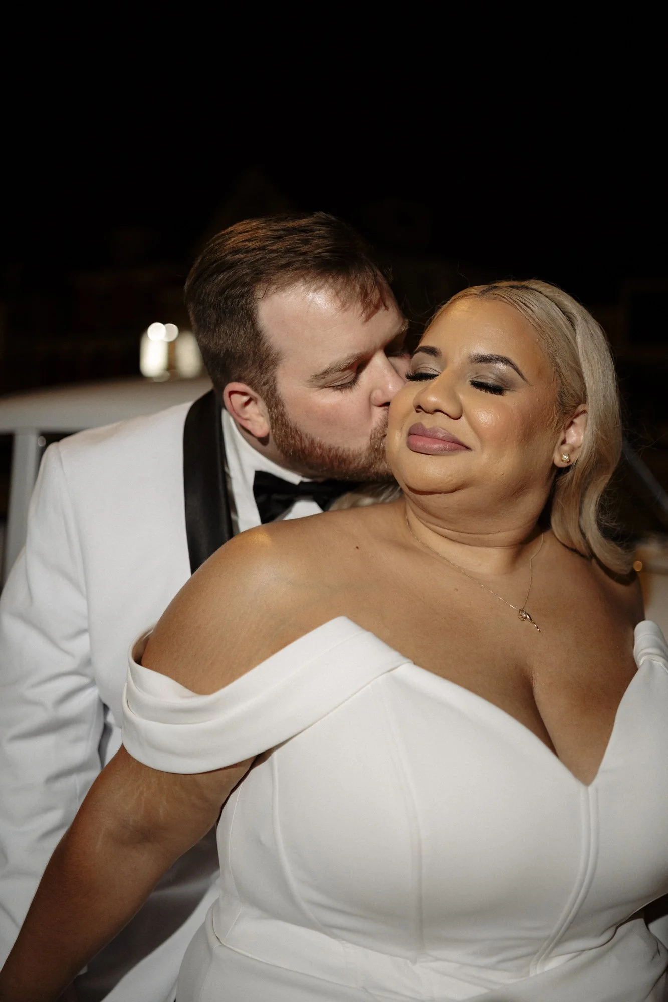 A man in a tuxedo is kissing a woman in a white wedding dress on the cheek, both with their eyes closed, at a wedding reception.