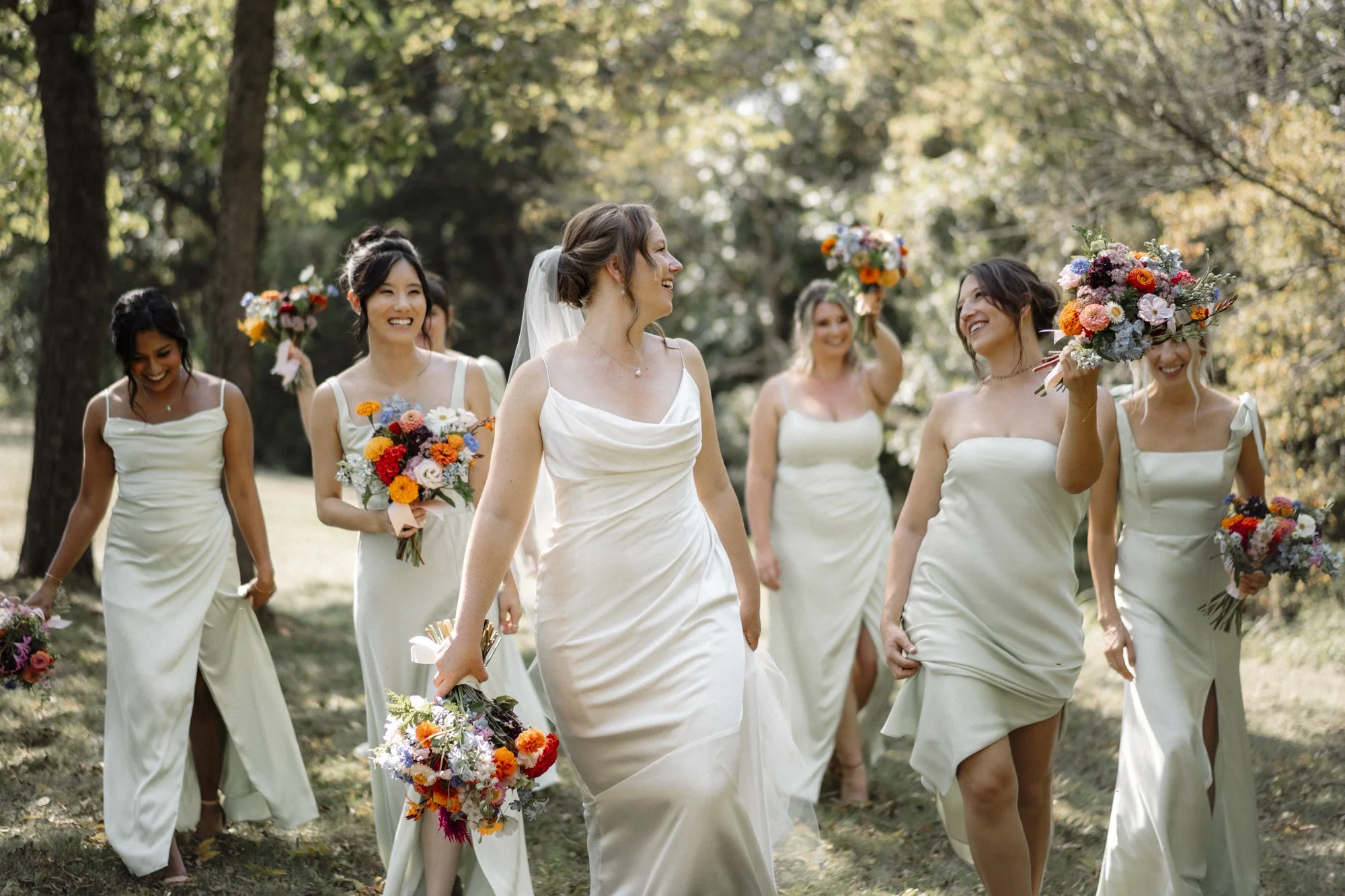 A bride and her bridesmaids walking outdoors in a forested area during daytime, holding bouquets of colorful flowers, smiling and enjoying the moment.