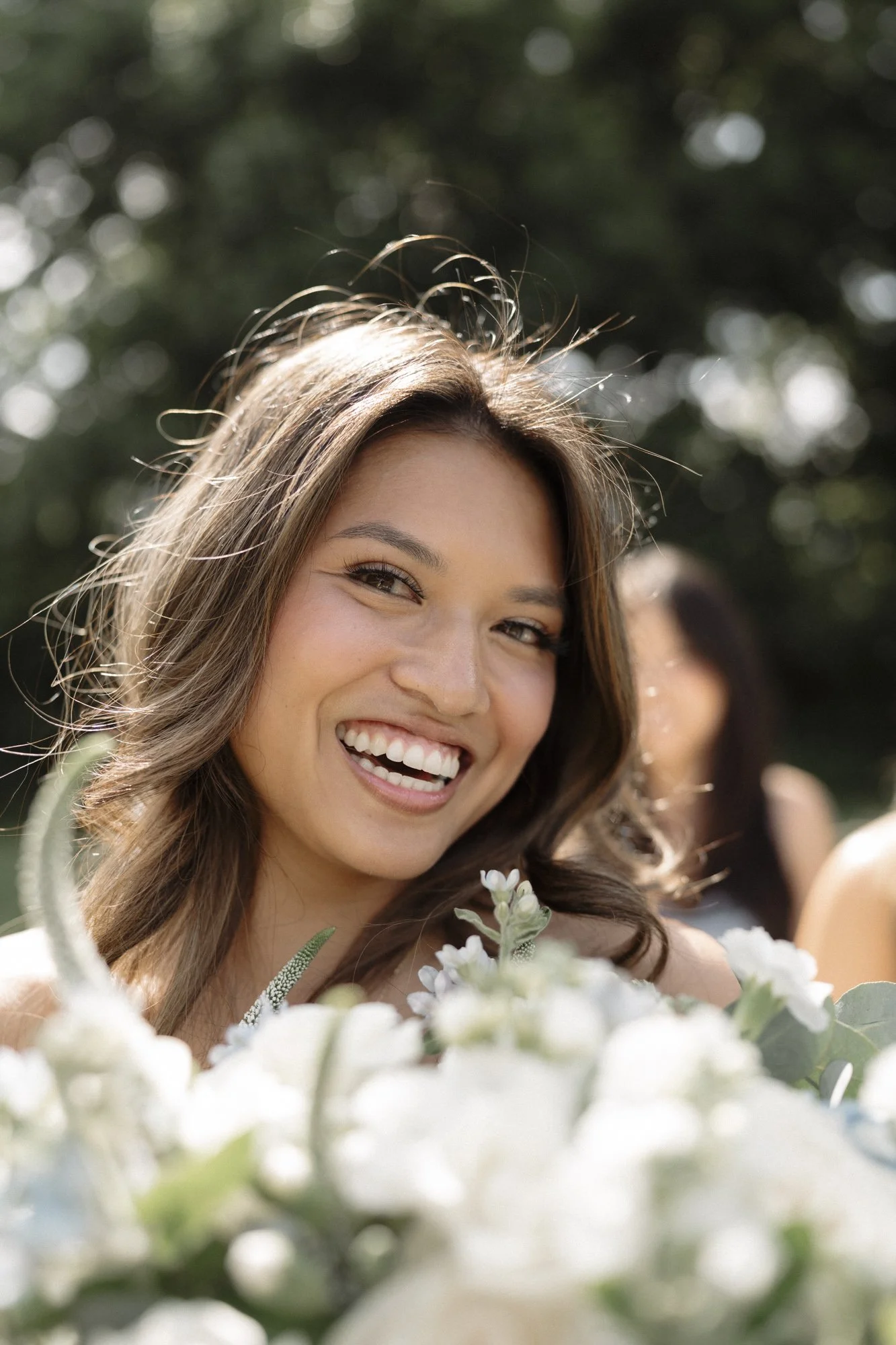Close-up of a smiling woman with long brown hair and light makeup, holding white flowers, outdoors with a blurred green background.