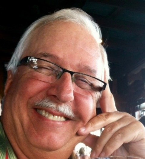 Close-up of a smiling older man with white hair, glasses, and a mustache, touching his face with his hand.