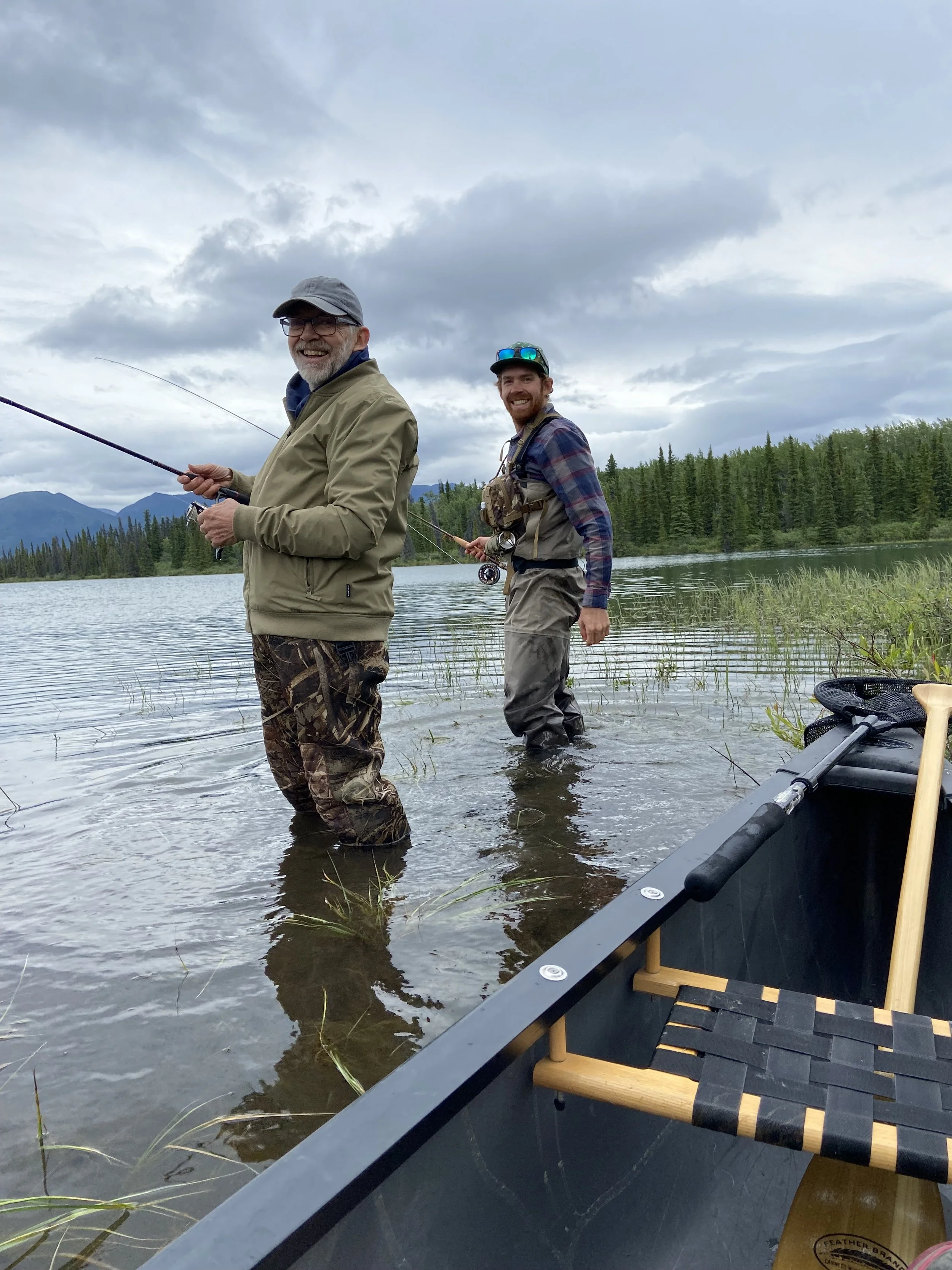 Two men fishing in a lake with mountains and trees in the background. One man is smiling and holding a fishing rod, while the other, standing behind him in the water, is also smiling and holding a fishing rod. There is a boat or kayak with fishing gear in the foreground.