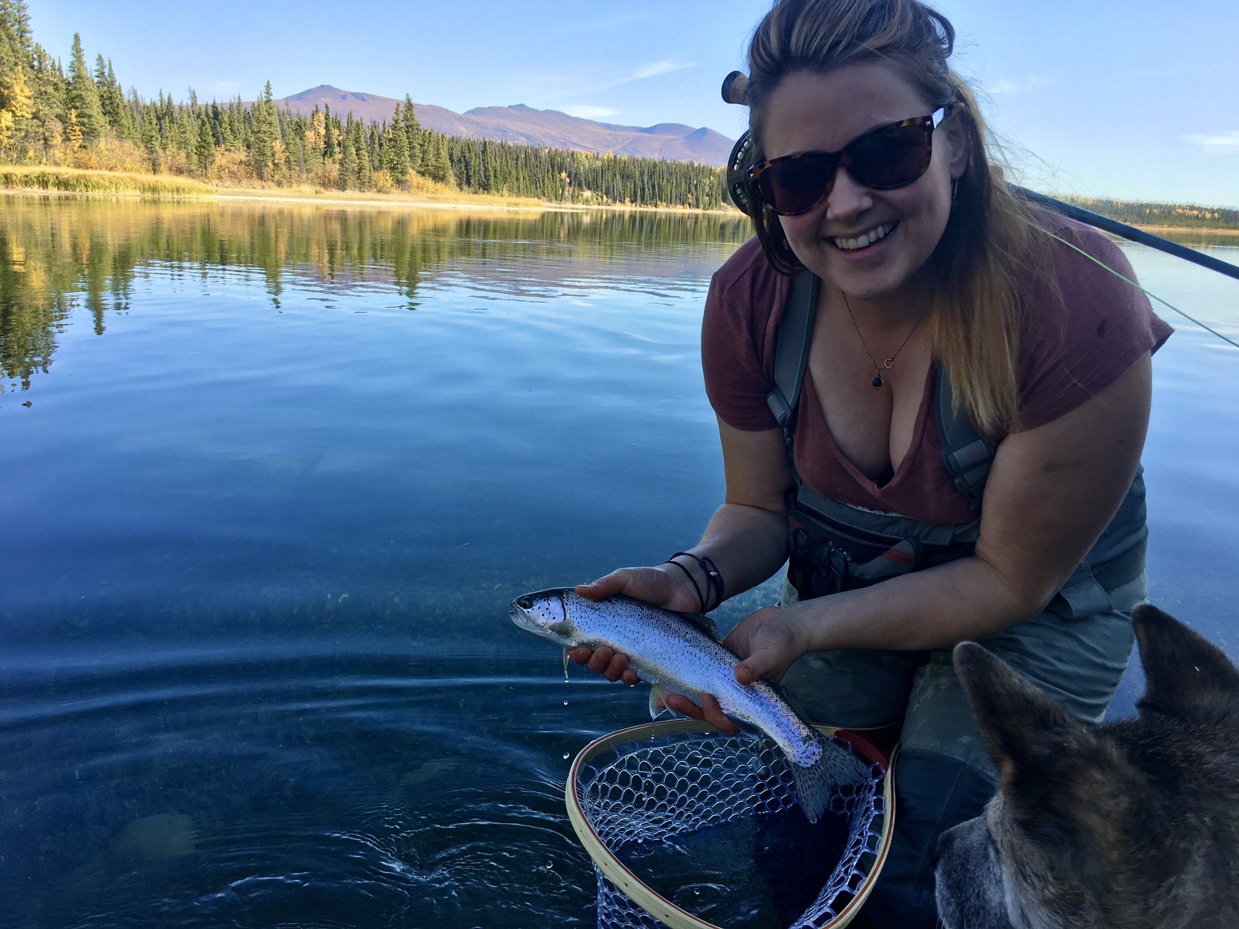 A woman smiling and wearing sunglasses, holding a fish over a body of water with a mountainous and forested landscape in the background.
