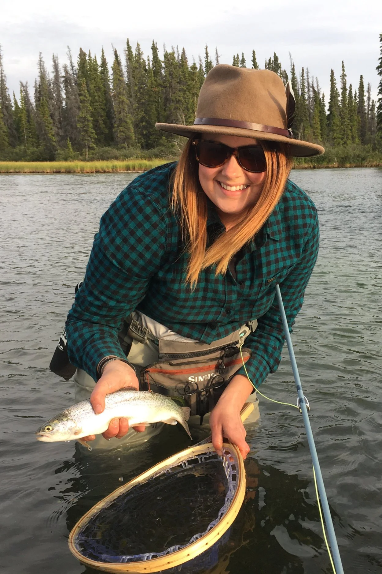 A woman in outdoor gear holding a fish she caught while standing in a river, with a fishing net in her hand, wearing a wide-brimmed hat, sunglasses, and a plaid shirt, with a backdrop of trees and water.