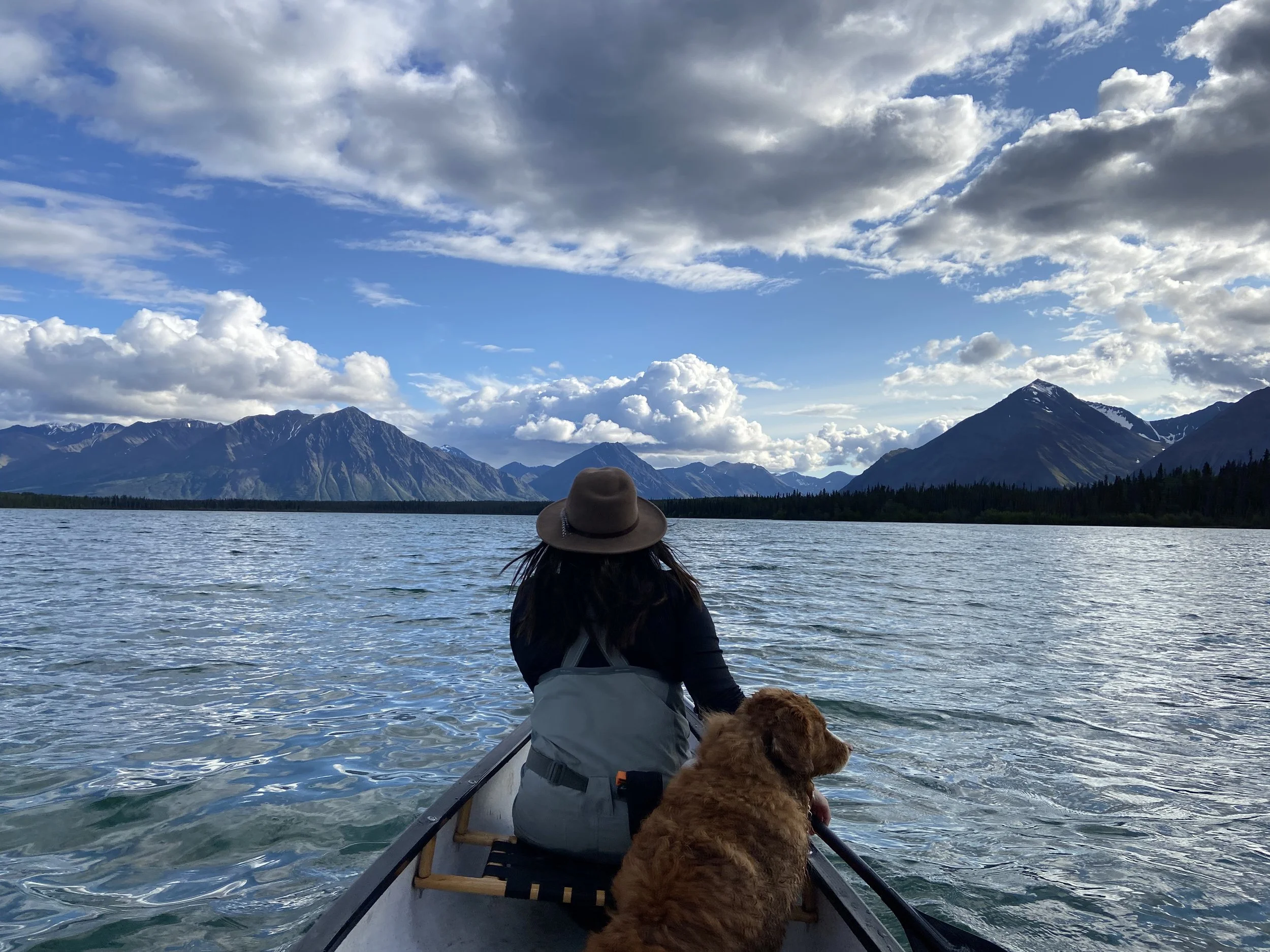 A person in a jacket and hat sitting with their dog in a canoe on a lake, with mountains and a partly cloudy sky in the background.