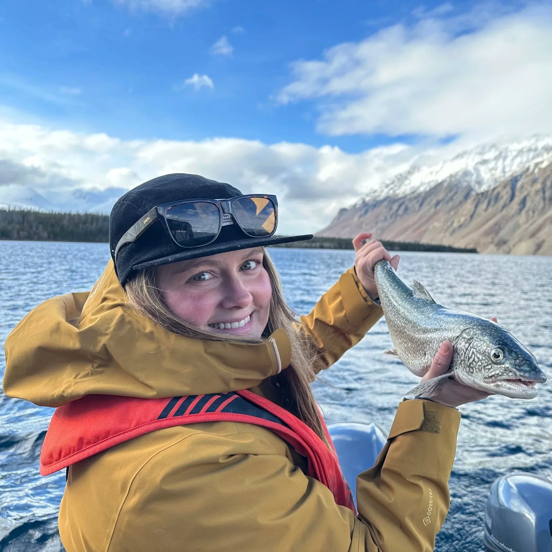 A woman holding a fish on a boat in a lake with snow-capped mountains and a partly cloudy sky in the background.
